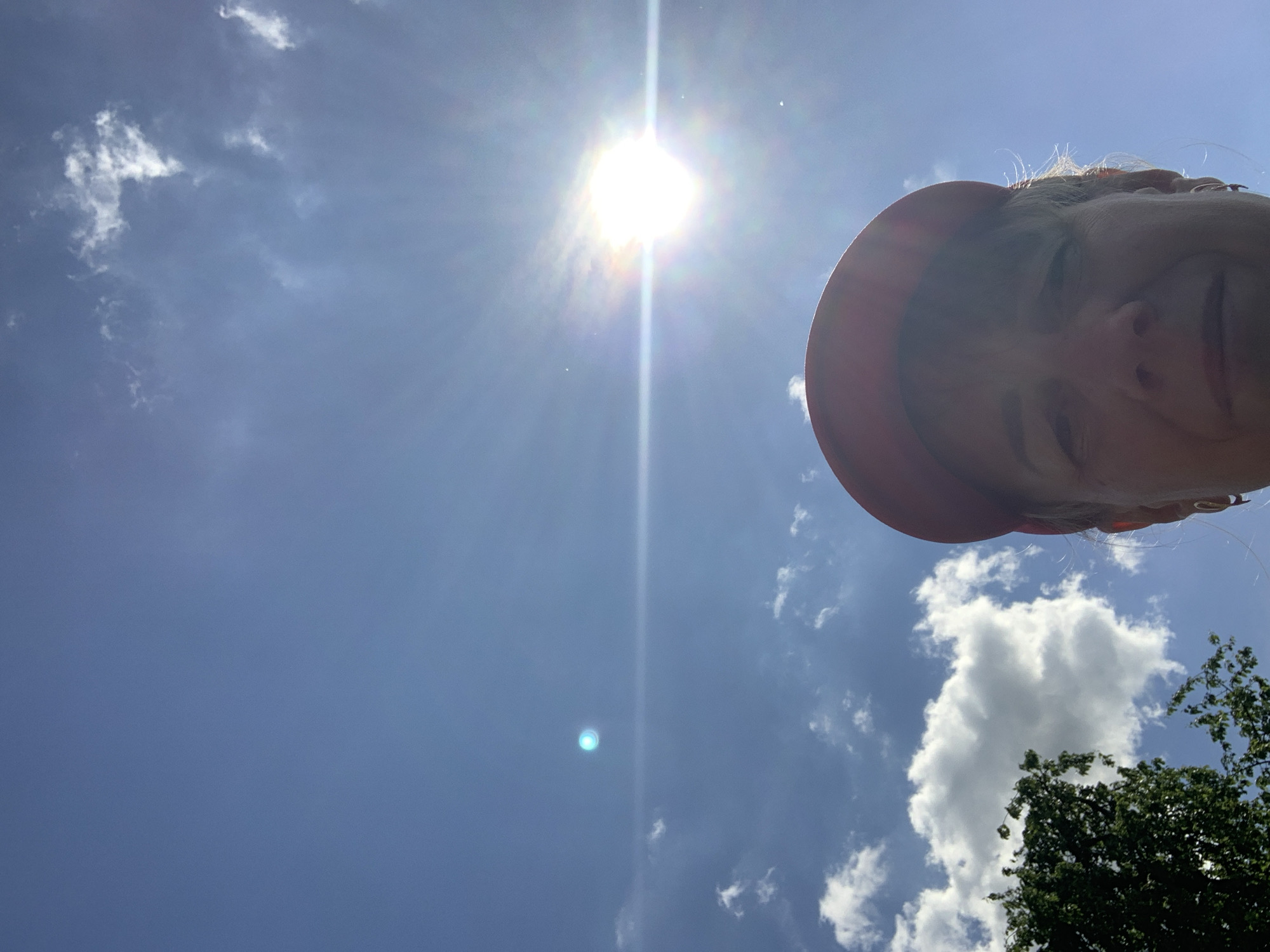 A low-angle, close-up shot shows the face of a person wearing a red baseball cap against a bright blue sky with sunbeams and scattered white clouds. A portion of a leafy green tree is visible in the lower left quadrant of the image. The sun is prominently displayed in the upper right quadrant, with a bright glare obscuring some of the details of the sky. The person's face is partially obscured by the shadow of the cap and the glare of the sun. There is a small, bright blue light anomaly in the sky to the left of the sun. The image appears to be taken outdoors. The location is Блеўчыцкі сельскі Савет, Belarus.