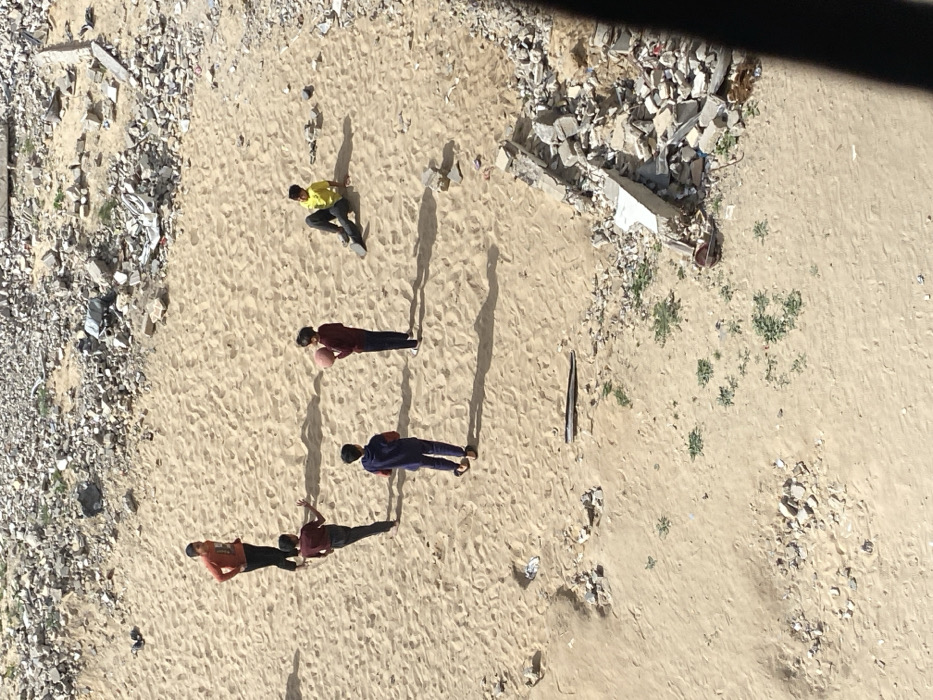 An overhead, high-angle shot captures five individuals, likely young males, engaged in activity on a sandy, open ground. The scene is dominated by the light brown sand, which exhibits ripples and texture, suggesting it's an unpaved, possibly urban or semi-urban area. Along the left edge and in the upper right portion of the frame, there are piles of rubble, broken concrete, and various debris, indicating a construction site, demolition area, or neglected ground. Sparse green vegetation sprouts in patches across the sandy expanse, particularly on the right side.
Five figures are visible, scattered across the sandy ground. One person, wearing a bright yellow t-shirt and dark trousers, is positioned in the upper-middle left, kneeling or crouching low to the ground. Directly below this individual, another person in a dark red or maroon top and dark trousers stands, holding a basketball in both hands. Further down and slightly to the right, a third person in a dark blue or purple top and dark trousers stands upright. Towards the bottom left of the frame, two more individuals are seen: one in an orange top and dark trousers, standing slightly ahead of another in a maroon or dark red top and dark trousers. All figures cast long, distinct shadows that stretch towards the right and slightly downwards in the frame, indicating a low sun angle, characteristic of either early morning or late afternoon. The presence of the basketball and the grouping of the individuals suggest they are playing or about to play a game on the sandy surface. A dark, elongated object, possibly a piece of wood or metal, lies on the sand to the right of the central figures. The top right corner of the image shows a dark, blurry edge, likely the structure from which the photograph was taken.
