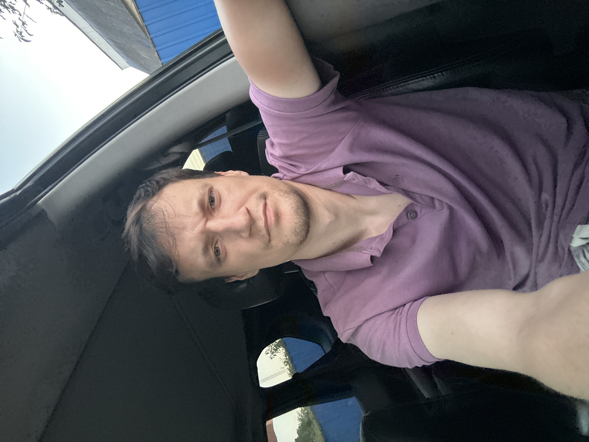 A man with short brown hair and stubble is sitting in the driver's seat of a car, looking towards the camera. He is wearing a purple polo shirt. The car's interior is dark, with black seats visible. Outside the car, through the windows, a blue structure and some trees can be seen against a bright sky. The man's left arm is resting on the car door. The image appears to have been taken during the day. There is no specific information available about the exact location in Shatava, Ukraine.