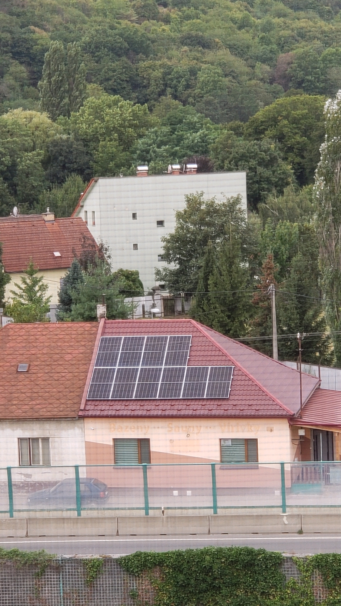 The image shows a residential area with houses and trees, set against a forested hillside. In the foreground, a building with a red tiled roof is visible, featuring an array of solar panels. Another building, lighter in color, is situated behind it. The scene is framed by a highway barrier and lush green vegetation along the road. The text "Bazený Souny Virivky" is visible on the facade of the building with solar panels, indicating it might be a commercial establishment related to swimming pools and saunas. The location appears to be in or near the city of Prešov, Slovakia. There are no people or animals visible in the image.