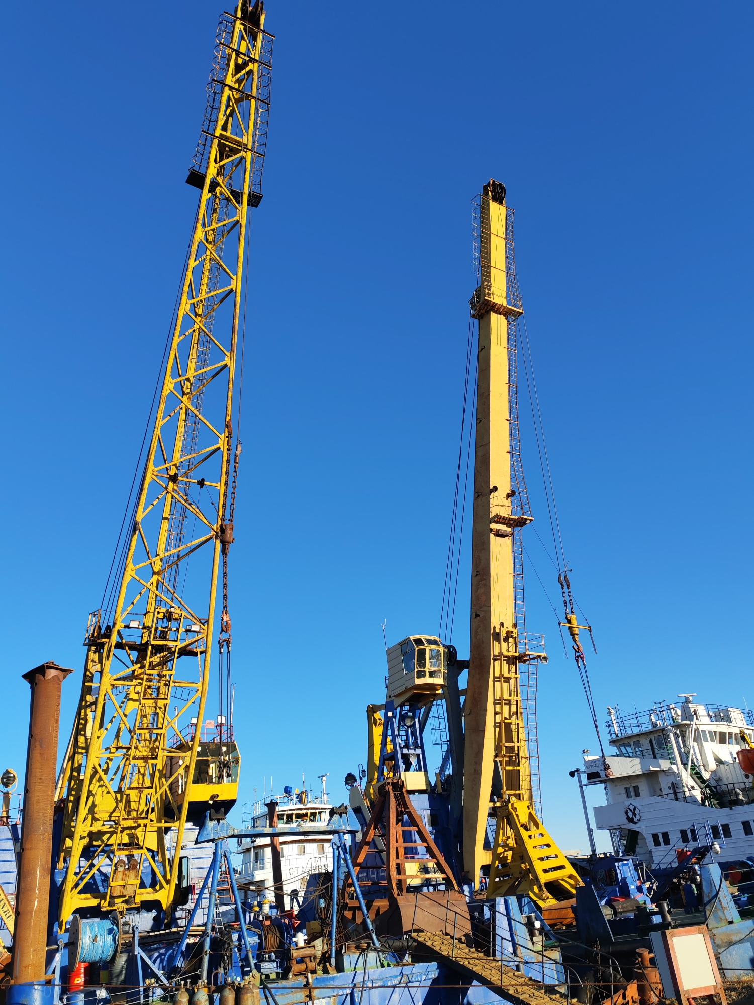 The image depicts a bright, clear day at a marine industrial site, showcasing two large yellow heavy-duty cranes or specialized machinery. On the left, a lattice boom crane with an operator's cab extends high into the cloudless blue sky, equipped with various cables and chains. To its right, a second tall yellow structure, possibly a pile driver or a different type of mast crane, also features an operator's cab and associated rigging.
These machines are mounted on what appears to be a blue-hulled working vessel or barge, with numerous industrial elements visible on its deck, including large pipes, winches, and metal structures. The word "PLAVUCHIY" (Russian for "floating" or "pontoon") is clearly visible in white letters on the side of this blue vessel, confirming its maritime nature. Partially visible in the background is a white ship or tugboat with a multi-deck superstructure.
The brilliant, unblemished blue sky indicates excellent weather conditions, and the strong, direct sunlight suggests it is mid-morning or early afternoon. No people are visible, emphasizing the scale and power of the industrial equipment. The scene conveys an atmosphere of heavy marine construction, dredging, or other large-scale offshore work. Faint text "NY.RU" is also discernible on a pipe on the far left.