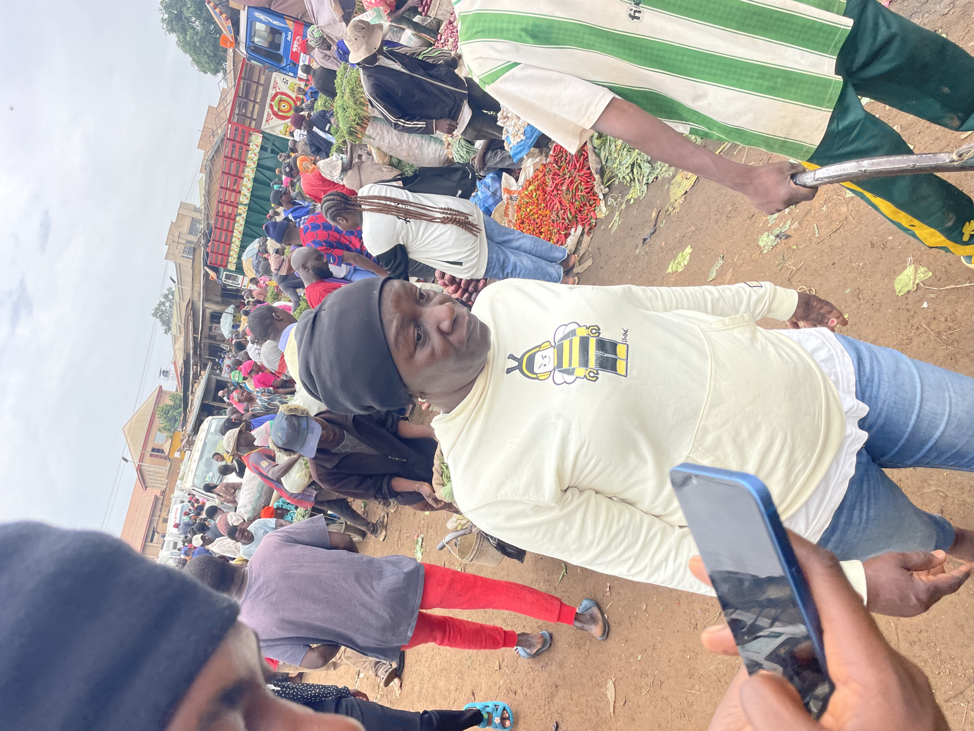 The image depicts a bustling outdoor market scene, likely in Gura Top, Nigeria. A crowd of people are gathered, engaged in various activities related to commerce. In the foreground, a person wearing a light-colored hooded sweatshirt with a bee graphic is looking to the right, holding a dark blue smartphone. Behind them, another person in a white and green striped shirt is visible, with their arm extended, holding what appears to be a bicycle handlebar or similar object.
The market is filled with stalls or displays of produce, including piles of red chili peppers and what seem to be vegetables like carrots or leafy greens. Several trucks and vans are present, suggesting the transport of goods. People are dressed in casual clothing, with a variety of colors and styles. Some individuals are wearing head coverings, common in this region. The ground appears to be dirt or packed earth, typical of an outdoor market.
The overall atmosphere seems lively and busy, characteristic of a market day. The sky is overcast, indicating a cloudy day, and the lighting suggests it is daytime. Although the exact time is not specified, the activity level implies it's likely during peak market hours. There is a sense of community and daily commerce in full swing. No significant text is clearly legible within the image.