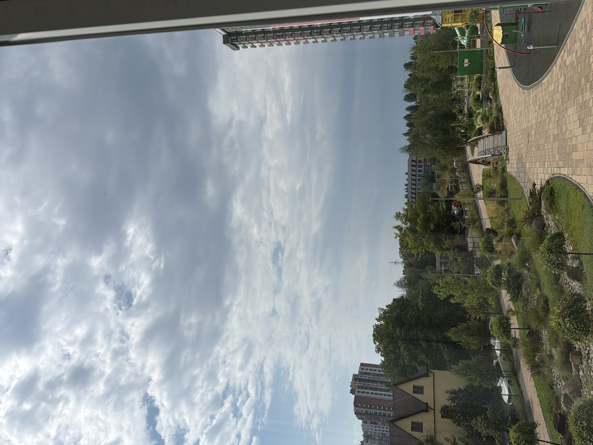 The image captures a cloudy sky over a residential area in Kyiv, Ukraine. In the foreground, a paved walkway curves around a playground with colorful structures and a small wooden bridge. Lush green trees and manicured landscaping are visible throughout the scene. Several apartment buildings, including a tall, modern high-rise with red accents, are scattered among the trees. The environment appears to be a well-maintained urban park or residential complex. No people are clearly visible in the image.