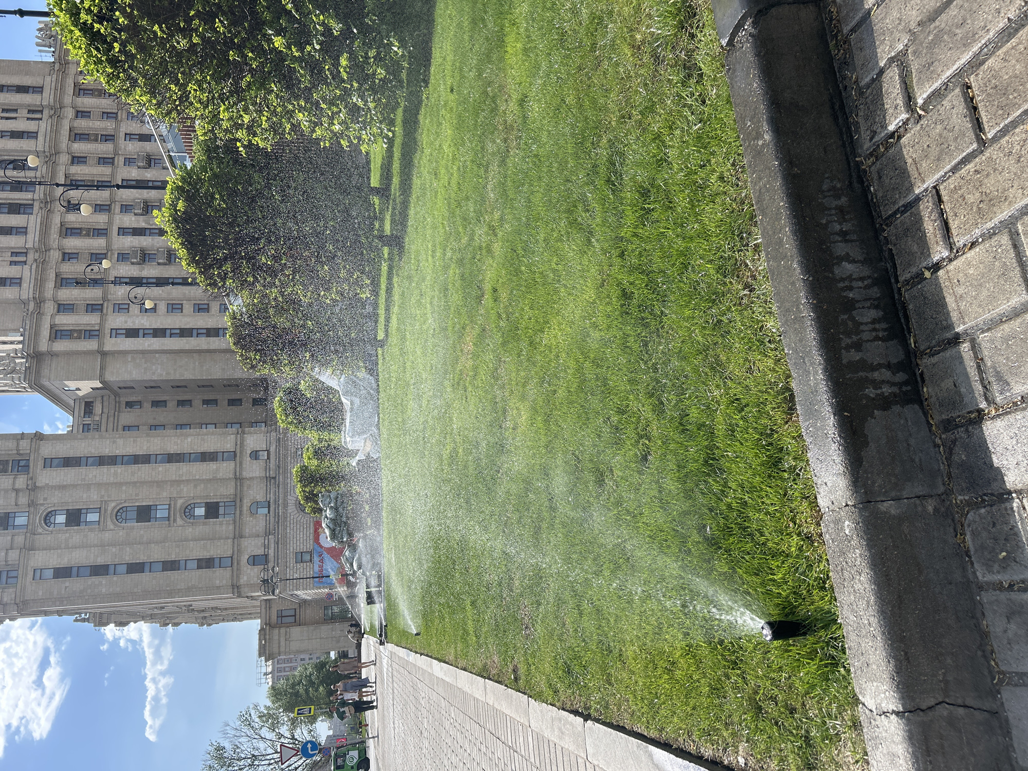 The image shows a grassy area being watered by sprinklers. The location appears to be an urban environment, based on the presence of a tall, multi-story building in the background. The building has a classical architectural style, featuring numerous windows and decorative elements. In the foreground, there is a sidewalk made of paving stones. A curb separates the sidewalk from the grassy area. The sprinklers are spaced along the edge of the grass, near the sidewalk. They are actively spraying water onto the grass, creating a misty effect. People can be seen walking along the sidewalk. The sky is blue with scattered clouds.