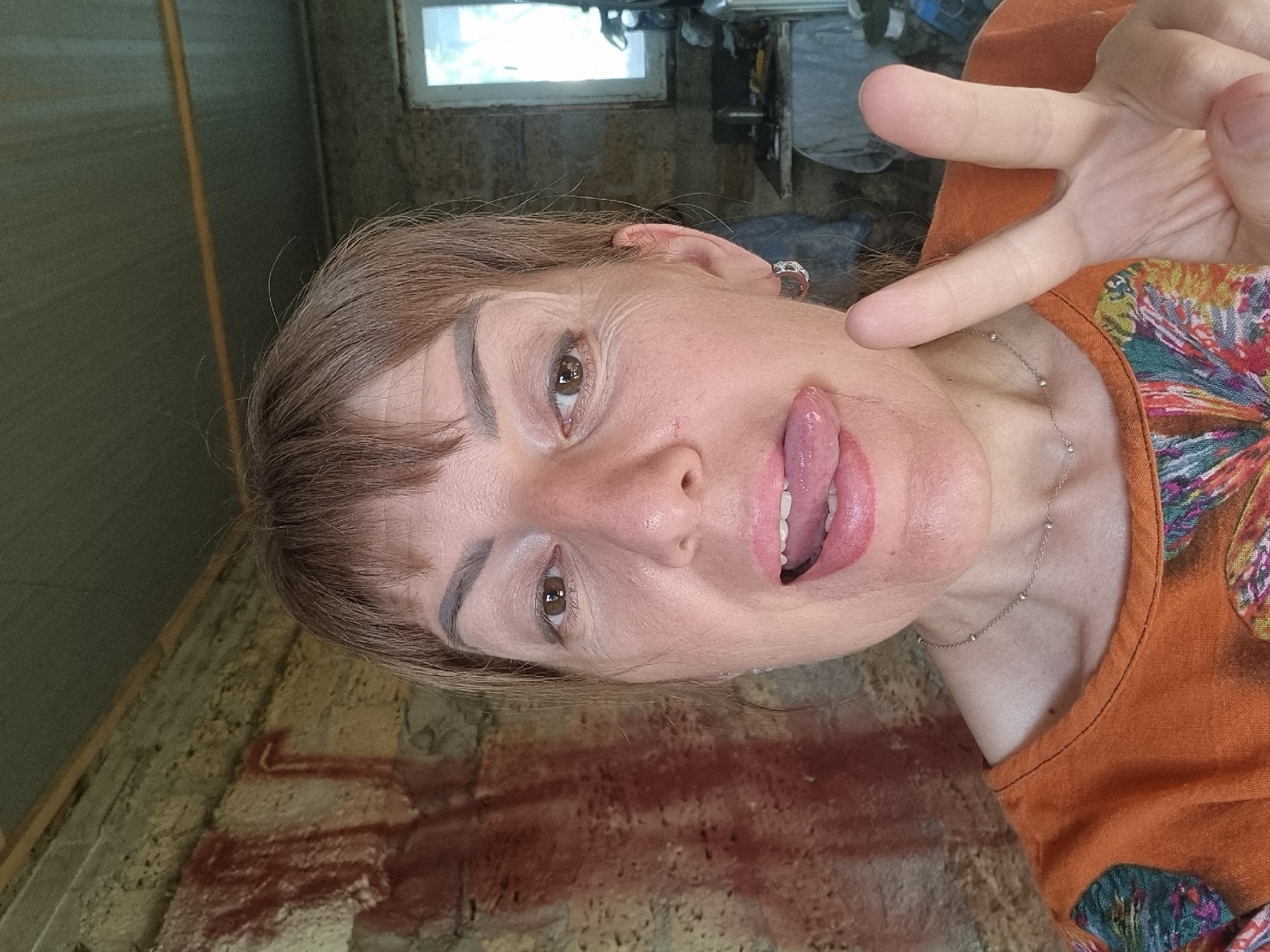 A woman is taking a selfie in what appears to be a dimly lit, rustic interior. The background is a rough, textured wall with red markings, possibly graffiti or paint splatters. To the right, a window is visible, showing daylight and what looks like another room or structure beyond. The woman is wearing an orange shirt with a colorful, patterned neckline. She is sticking out her tongue and making a peace sign with her right hand. Her hair is light brown and cut in a short, fringed style. She has on hoop earrings. The image is a close-up of her face and upper body, suggesting a casual and playful mood. There is no discernible information about the specific location within the city of Blizhnee, Ukraine.
