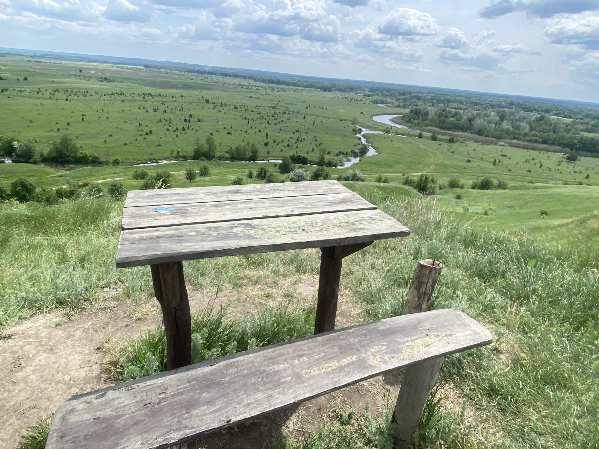 The image depicts a rustic picnic table and bench situated on a grassy hillside overlooking a broad, green valley. A winding river cuts through the valley floor. The picnic table is constructed of weathered wood and appears worn. The valley is predominantly grassland interspersed with scattered trees and shrubs. The sky is partly cloudy. The location is Ustyvytsia, Ukraine.