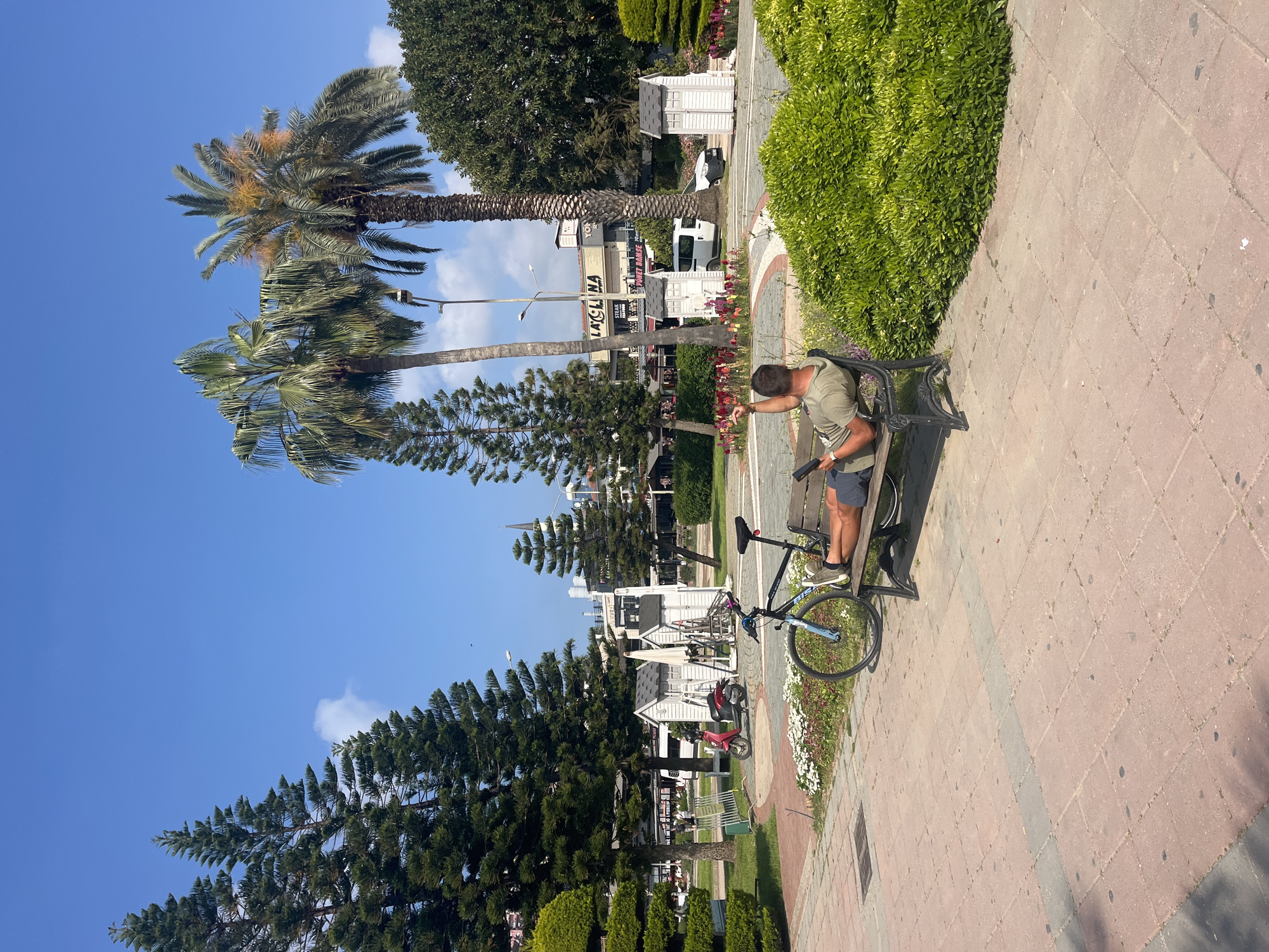 The image displays a sunny outdoor scene, likely a park or promenade in Alanya, Turkey. A man is seated on a black, ornate metal park bench on the right side of the frame. He is dressed in a dark green short-sleeved shirt and dark shorts, with a backpack slung over his shoulder, leaning forward and pointing downwards towards the ground. A black and light blue bicycle is parked next to the bench.
The foreground features reddish-brown paved pathways, bordered by manicured green hedges and flower beds containing white and red blossoms. The scene is lush with vegetation, including several tall palm trees with visible trunks and fronds, as well as dense, conical evergreen trees.
In the mid-ground, beyond the trees and pathway, are multiple small, white, house-like structures, possibly kiosks. Further in the background, various buildings are visible. Text on one building clearly reads "LAGUNA," with "STEAK HOUSE" and partially visible words like "BAR" or "BEER" below it on adjacent signs. A white van and a small red moped are also parked in the background.
The sky is clear and blue with scattered white clouds, indicating bright daylight, likely late morning or early afternoon. The weather appears pleasant and sunny. The man's posture suggests a moment of rest or adjustment during an activity, possibly cycling.