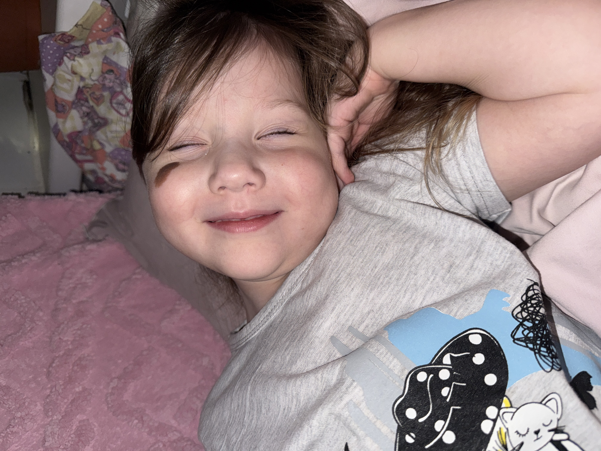 A young child lies on a pink bed with their eyes closed, smiling slightly. They are wearing a gray shirt with a cartoon design featuring a white cat and other black and white elements. Their hand is raised near their head. A patchwork-like fabric is visible in the upper left corner of the frame. The location is unknown and unrelated to Кипчак, Russia.