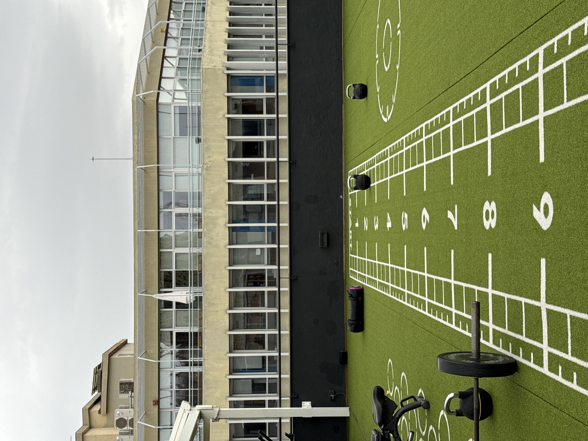 The image shows a rooftop gym area in Barcelona, Spain. The foreground displays green artificial turf marked with white lines and numbers resembling a workout grid. Various gym equipment is scattered on the turf including kettlebells and a barbell loaded with plates. Adjacent to the workout area is a tall building with a facade that is mostly glass windows set in a pale yellow or beige frame. The building is multiple stories high and features metal railings and scaffolding along its exterior. The sky is overcast and gray, suggesting a cloudy day.