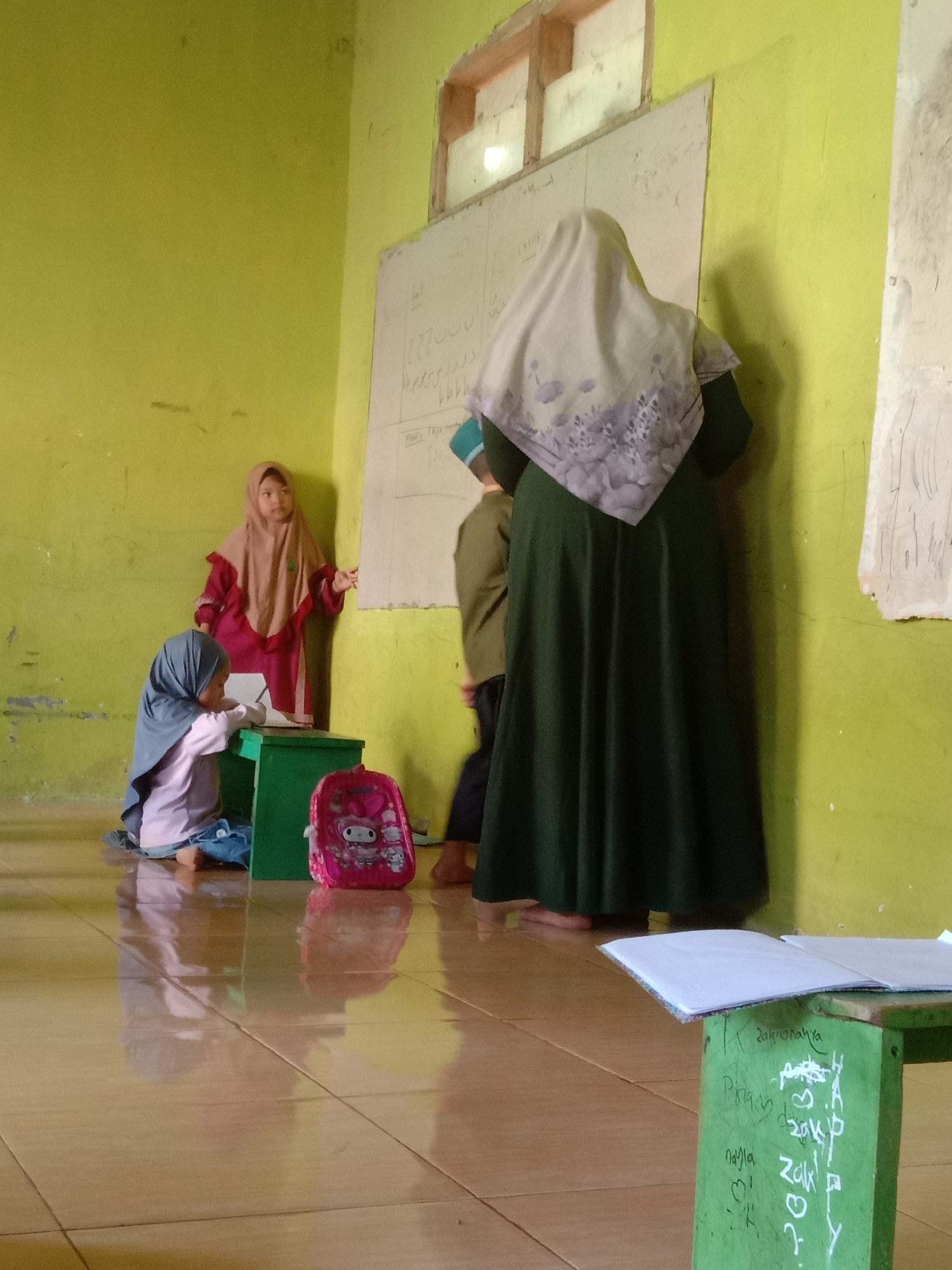 In a classroom in Tulungrejo, Indonesia, a teacher in a green dress and a headscarf stands facing a whiteboard, her back to the camera. A young girl in a pink and beige hijab is standing to the left, near the whiteboard, pointing at it. Another young girl, wearing a grey hijab and a light purple top, sits on the floor to the left of the standing girl, looking down at a green box. A pink backpack with a character design sits on the floor next to the sitting girl. The room has light green walls and a tiled floor that reflects the light. There is also a green table in the foreground with papers on it, and writing on the side of the table. The whiteboard has some writing and drawings on it. Another child, partially visible and wearing a brown jacket, stands between the teacher and the standing girl.