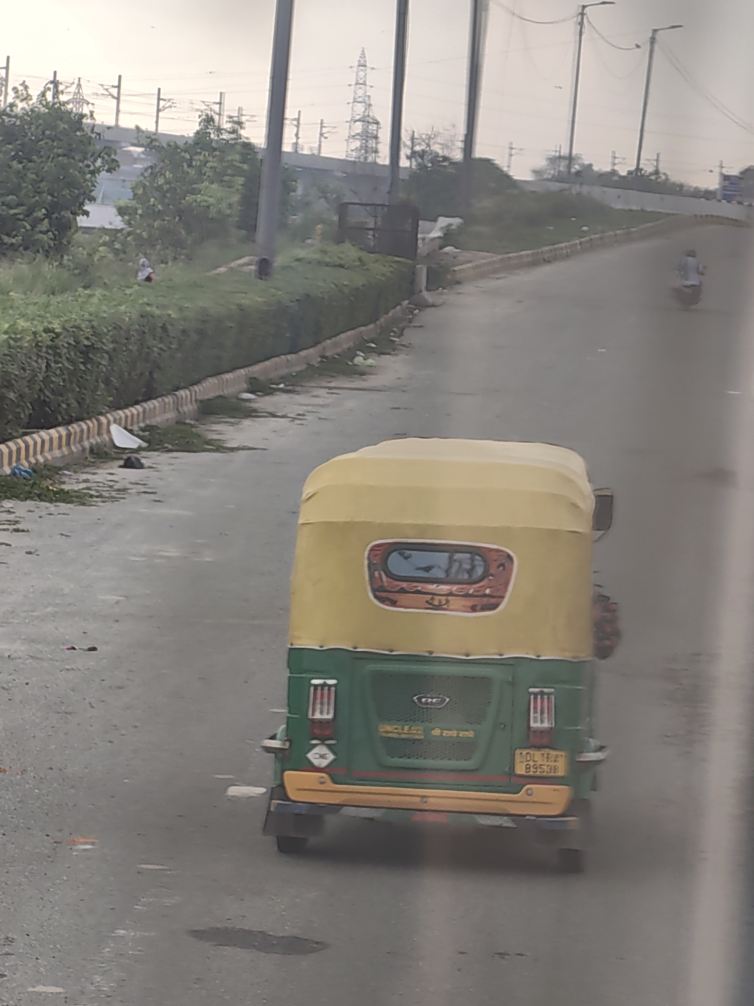 The image depicts an outdoor scene on a road, likely in Delhi, India, given the presence of an auto-rickshaw with a Delhi registration plate. In the foreground, a green and yellow auto-rickshaw is the primary subject, viewed from behind. The auto-rickshaw has a yellow roof and a green body, with a yellow bumper. Its rear windshield features a decorative element with an image. A yellow license plate with the registration "DL 1RA 8953R" is visible on the back. To the left of the auto-rickshaw, a roadside area is lined with a concrete barrier that has alternating black and yellow stripes. Beyond the barrier, there is dense green foliage, and further back, a person in light-colored clothing is partially visible amidst the greenery. In the background, power lines and transmission towers suggest an urban or semi-urban setting. A motorcycle is also seen in the distance on the road. The overall scene appears to be captured during daylight, with the sky being overcast or hazy.