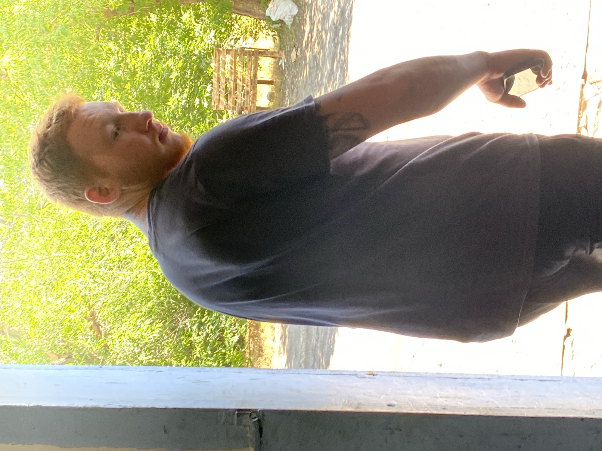 An adult Caucasian male with red hair and a beard is standing, facing away from the camera, with his head turned to the right. He is wearing a dark gray t-shirt and dark pants. He has a tattoo on his left arm. He is holding a dark object in his right hand. The man is standing in an open doorway, with a sunlit outdoor area visible beyond him. The outdoor area contains green foliage, a wooden bench, and a tree. The ground is dirt and concrete. The scene appears to be in an outdoor, possibly rural or industrial, setting.