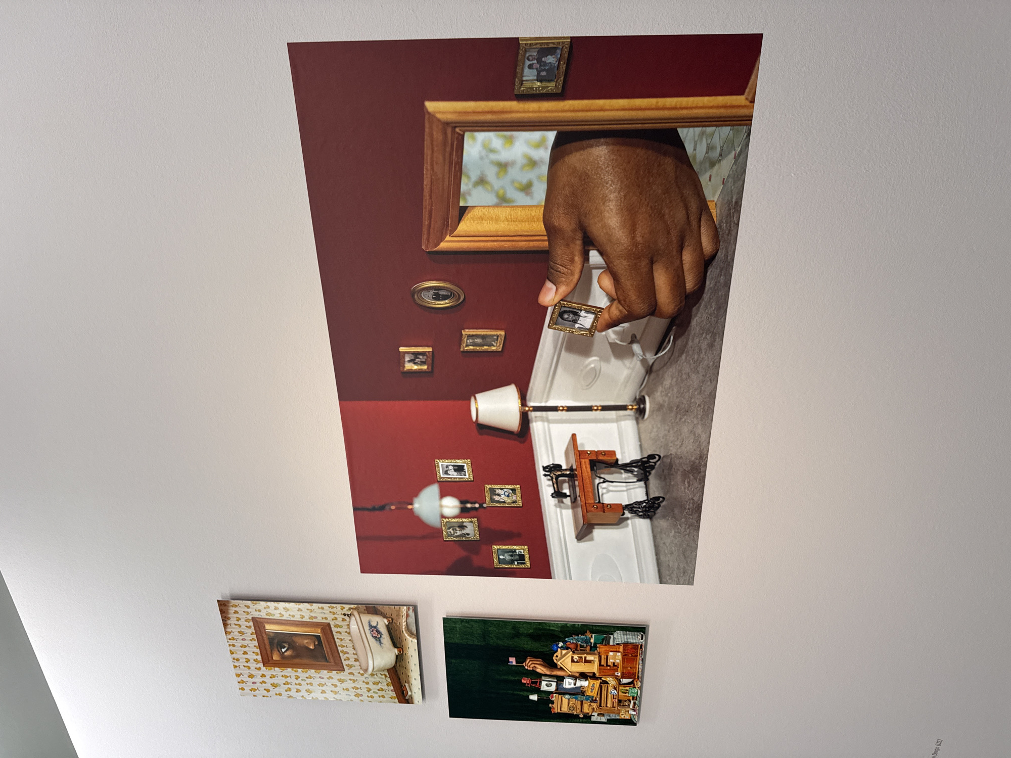 This image displays a wall in an exhibition space, possibly a gallery, in Frankfurt, Germany, as indicated by a partially visible text in the lower left corner. The wall is adorned with three framed artworks.
The largest artwork, positioned centrally and to the right, is a hyperrealistic, surrealist depiction of a room with a deep red wall. In the foreground, a miniature scene is rendered with meticulous detail. A small, antique-style sewing machine sits on a wooden table. A floor lamp with a white shade stands to the right of the table. The wall is decorated with four small, framed photographs. In the middle ground, a large, gilded picture frame appears to be a doorway, revealing a background with a yellow, bird-patterned wallpaper. A dark-skinned hand emerges from this doorway, holding a tiny framed photograph of a person. The hand is the focal point of this artwork, drawing attention to the act of holding and presenting the photograph.
To the left of the main artwork, and slightly higher, is a second framed piece depicting a close-up of a person's eye reflected in a bathtub, set against a patterned wallpaper. Below this, and to the left, is a third framed artwork, a vertical composition showcasing a miniature, densely packed arrangement of various objects, including what appear to be bottles, boxes, and buildings, with a hand holding a small American flag emerging from the arrangement.
The lighting in the exhibition space appears to be artificial, creating subtle shadows and highlighting the textures of the artworks and the wall. The wall itself is painted a neutral, off-white color.
