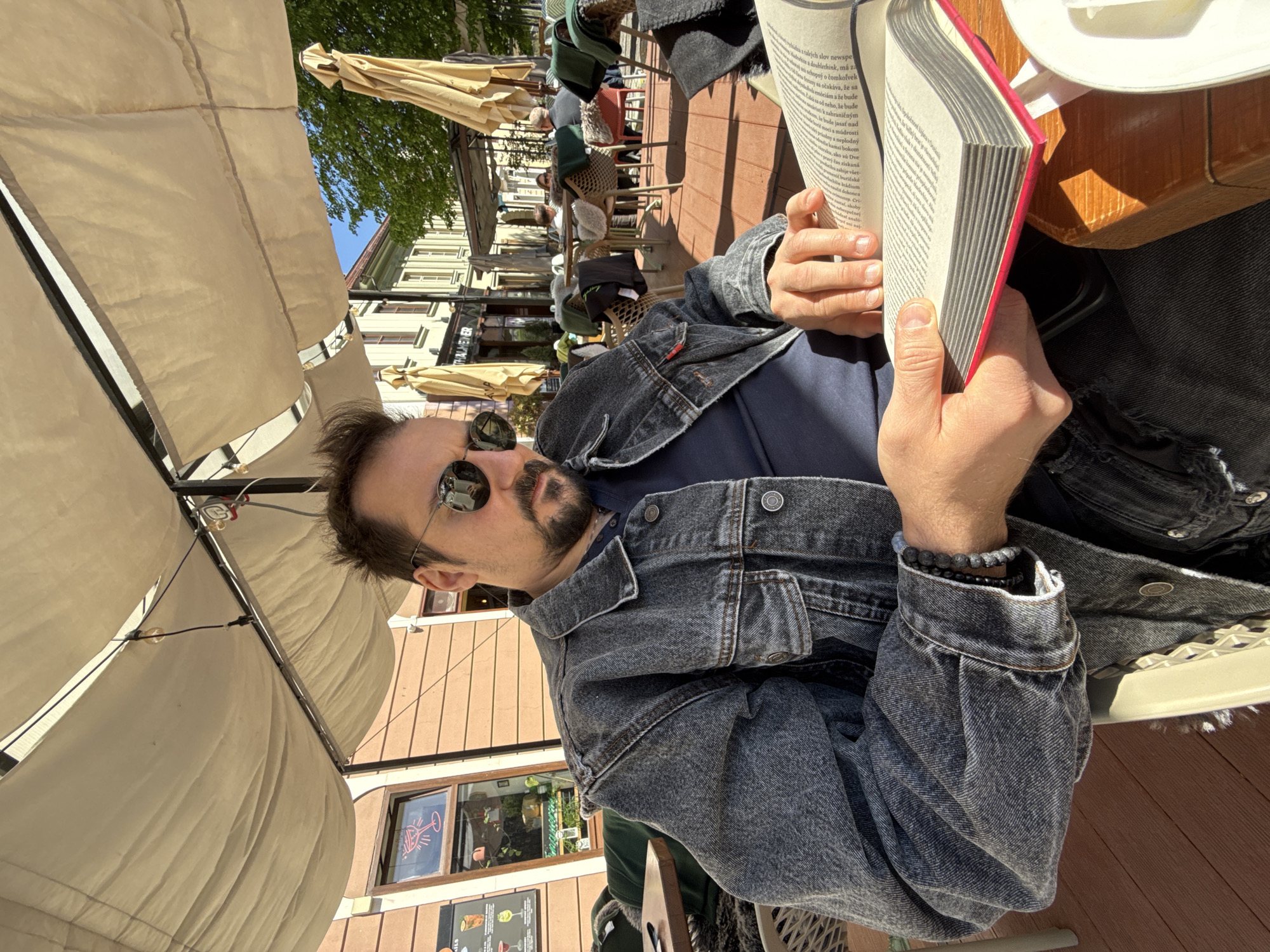 The image shows a man sitting at an outdoor cafe, likely in Trnava, Slovakia. He's wearing a denim jacket, a dark t-shirt, and sunglasses. He has a beard and is holding a book with red accents, apparently reading. There's a white mug or dish on the table next to the book. The cafe has outdoor seating under large beige umbrellas. Other patrons are visible in the background, along with buildings that appear to be part of the city's architecture. A restaurant named "Thalmeiner" is also partially visible in the background. The cafe is on a brick surface. The image is shot from an upward angle.