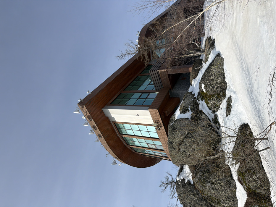 The image displays a multi-story, modern building situated on a snow-covered, rocky hillside in Zelenaya Polyana, Russia. The structure features a distinctive curved facade, clad primarily in brown wood paneling, with large, rectangular, turquoise-tinted windows dominating the primary visible section. The roof is flat and equipped with multiple white dish-shaped satellite antennas and other communication devices. Below the building, large grey boulders emerge from patches of white snow, which cover much of the ground. Sparse, bare deciduous trees with thin branches are visible throughout the landscape, lacking foliage. The sky above is clear and light blue. No individuals, activities, or interactions are present in the scene.