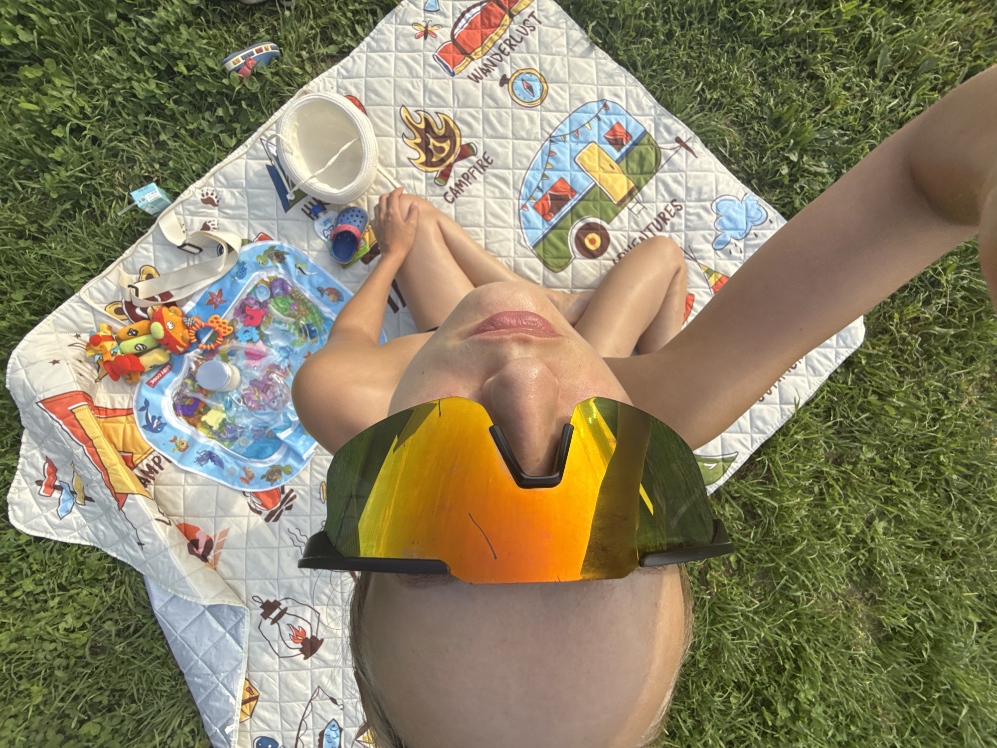 This overhead shot shows a person lying on a picnic blanket spread on green grass. The person is wearing large, mirrored sports sunglasses that reflect the sky and surrounding greenery. Their legs are crossed, and their arms are visible. The picnic blanket is patterned with camping-themed illustrations, including a camper van, a campfire, a compass, and lanterns, with the word "WANDERLUST" and "ADVENTURES" visible. To the left of the person, a small inflatable water playmat with toys is visible, along with a white straw hat and a pair of colorful flip-flops. The lighting suggests it is a sunny day, likely during daylight hours. The setting appears to be outdoors in a grassy area, possibly a park or backyard, in Подшивалово, Russia.