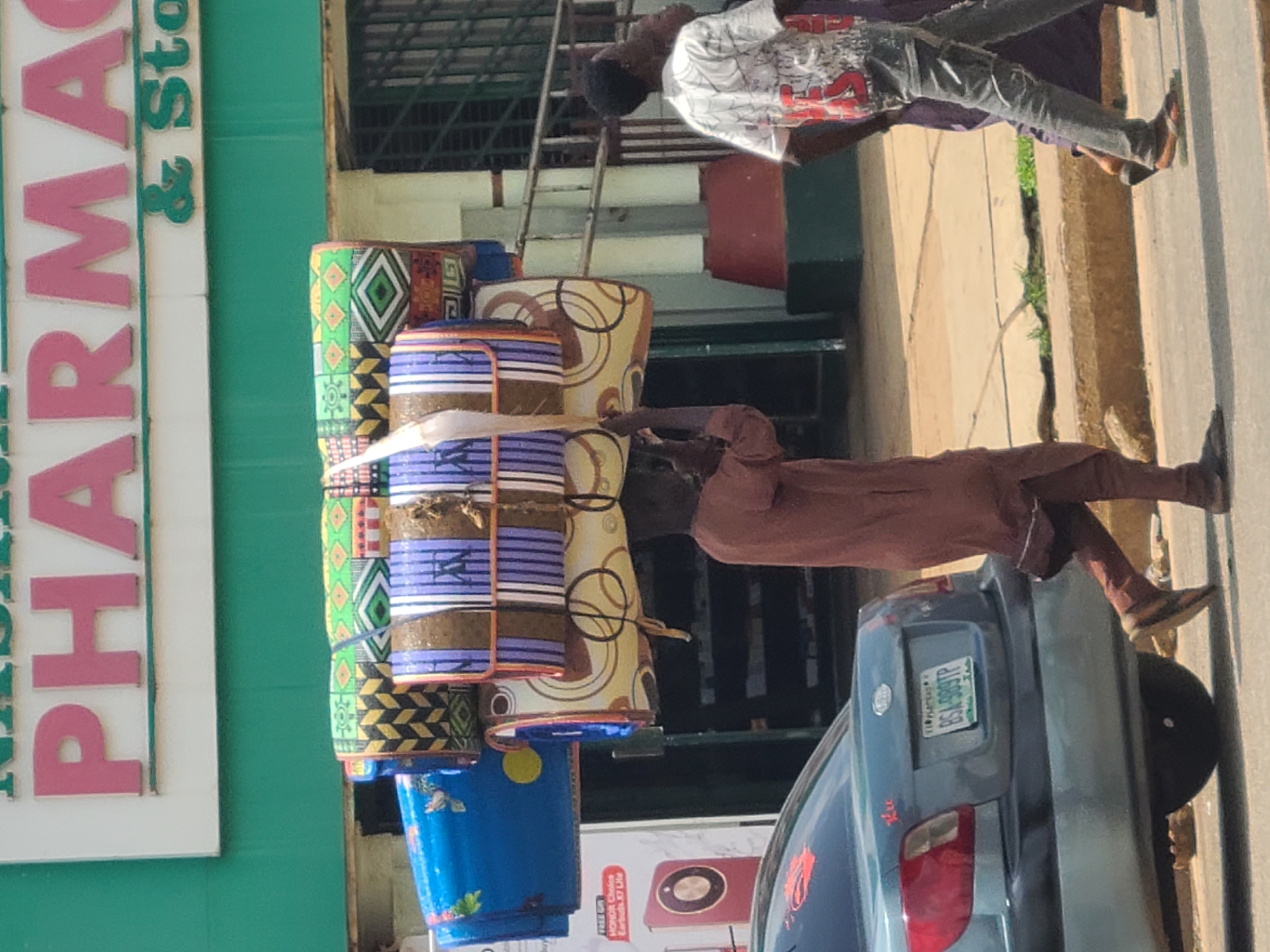In Jos, Nigeria, a man with dark skin and wearing a brown robe walks carrying a large bundle of rolled-up mats on his head. The mats are colorful, with geometric patterns in green, white, yellow, and purple. To the left of the man, a blue bucket-like object is visible. Behind the man, another person in a white t-shirt with red and black graphics and grey pants is walking, followed by another person partially obscured. A light blue car with a license plate reading "BS4-989TP" is parked on the left side of the street. A building with green and white signage is visible in the background, with the word "PHARMACY" in large pink letters. The scene appears to be on a street in a commercial area.
