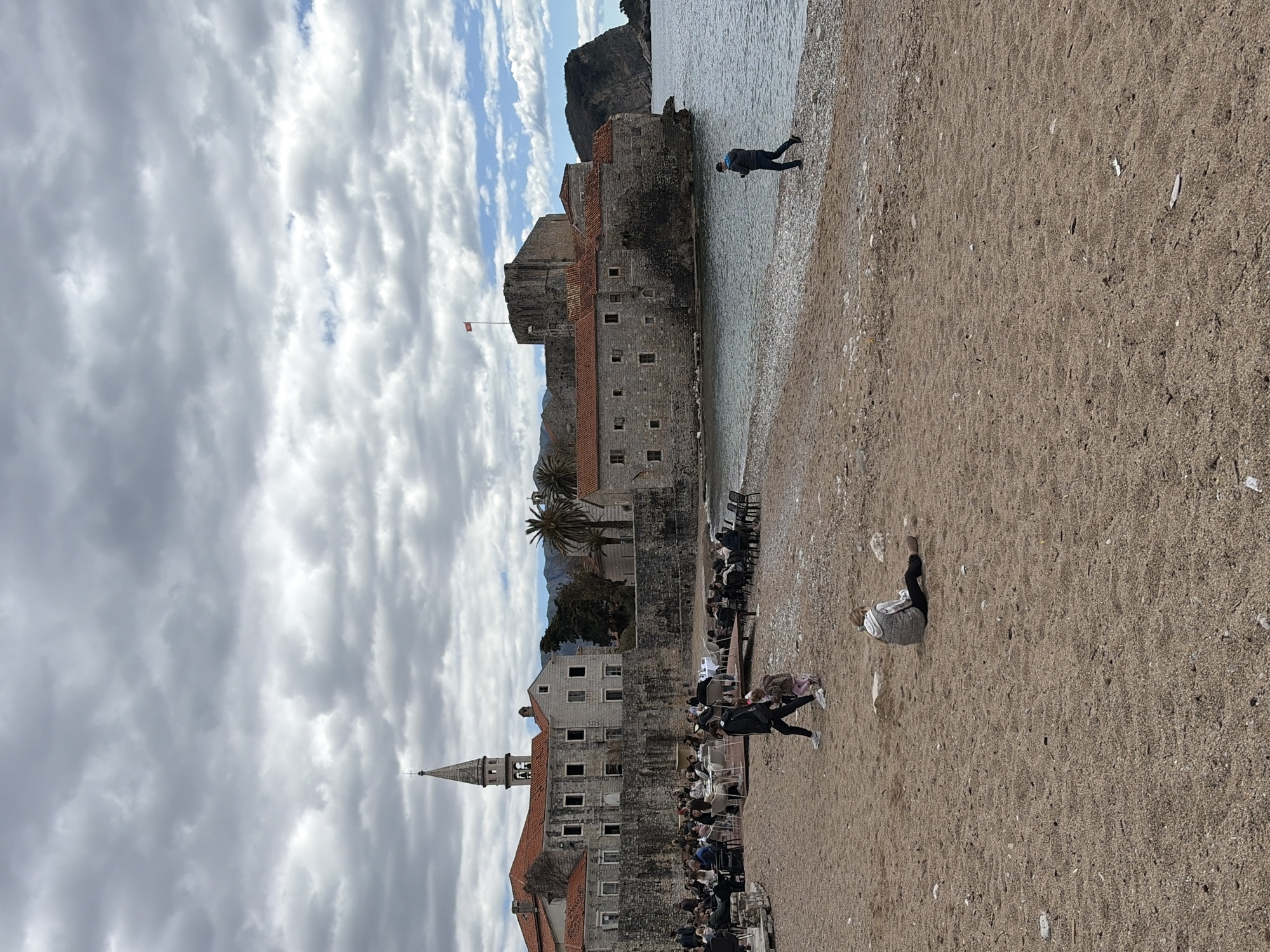 The image shows a view of the old town of Budva, Montenegro, from a beach. The old town features stone buildings with red tile roofs, including a church with a prominent steeple. A fortress stands closer to the sea, partially surrounded by water. A flag is flying from the fortress. People are seen on the beach. One individual is walking in the water. Others appear to be attending an event with rows of chairs. The sky is cloudy.