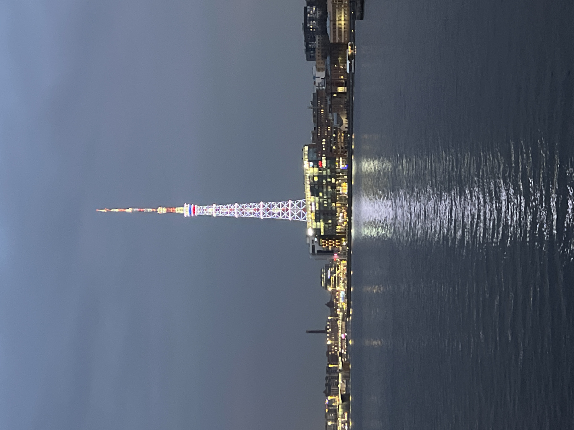 The image depicts a nighttime or deep twilight scene of the Neva River in Saint Petersburg, Russia, with the illuminated Saint Petersburg TV Tower as the central focus. The tower, a tall lattice-steel structure, stands prominently on the far bank, displaying red, white, and blue lights along its spire and white lights outlining its main framework.
On the river's far bank, a cityscape extends left and right of the tower. Buildings, a mix of modern and older architecture, are illuminated with various interior and exterior lights, predominantly warm yellow/orange, contributing to the ambient glow reflected in the water. To the left of the tower, a bridge with vehicle lights is visible spanning the river, and a tall, dark industrial chimney rises in the mid-distance. The immediate foreground is dominated by the dark, rippled surface of the Neva River, which reflects the bright lights of the tower and the city. The sky above is a uniform dark gray-blue, devoid of visible celestial bodies or distinct cloud formations. No individuals, specific actions, or interactions are observable within the frame.
