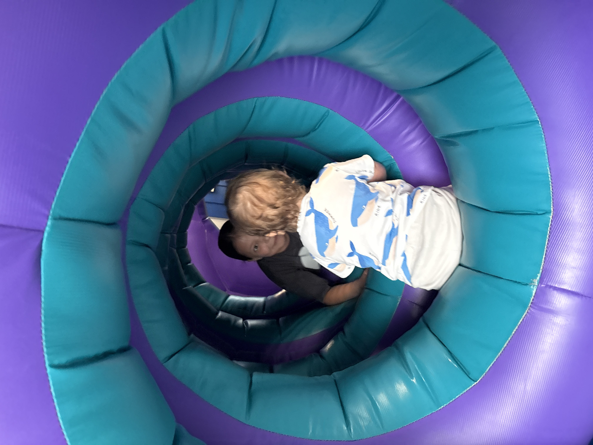 Two children are inside an inflatable spiral tunnel. The tunnel alternates between purple and turquoise rings. One child, with blond hair, wears a white shirt with blue whale patterns. The other child wears a dark gray shirt. The tunnel appears to be indoors. The location is not identifiable as Izhevsk, Russia, based on the image.
