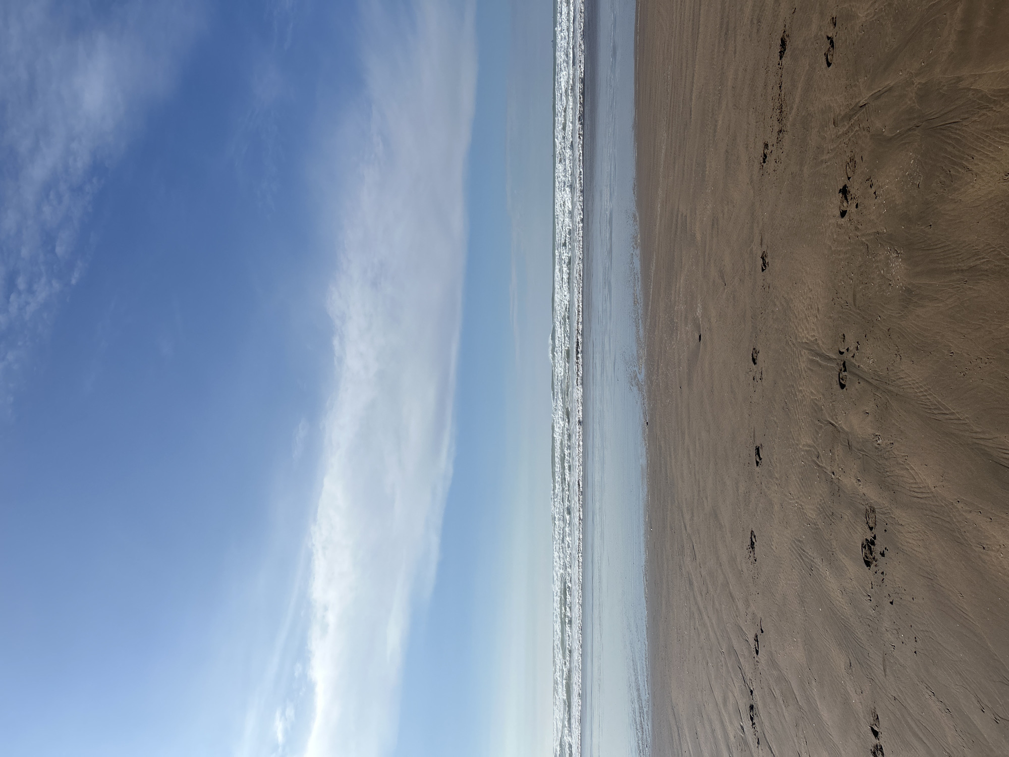 The image depicts a beach scene in Casablanca, Morocco. The sky occupies the left side of the frame, displaying a gradient of blue with scattered, wispy clouds. The right side shows the beach, with wet sand reflecting the sky, bordered by the ocean with small waves. Footprints are visible in the sand, indicating pedestrian activity. There are no people visible in the frame.