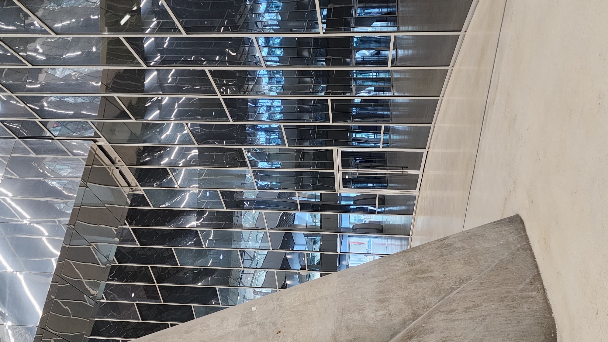 The image shows the interior of a modern building with a distinctive glass facade. A large, curved concrete structure dominates the left foreground, contrasting with the sleek, reflective surfaces of the building's exterior. The glass walls are divided into numerous rectangular panels, some of which are dark and reflective, while others show distorted reflections of the interior spaces and lighting fixtures. A set of double glass doors with silver frames is visible in the middle ground, providing an entrance into the building. The floor is a smooth, light-colored material, possibly polished concrete or stone. The overall impression is one of contemporary architecture, emphasizing light, reflection, and geometric forms. The image appears to be taken in the city of Moscow, Russia, suggesting a public or commercial building.