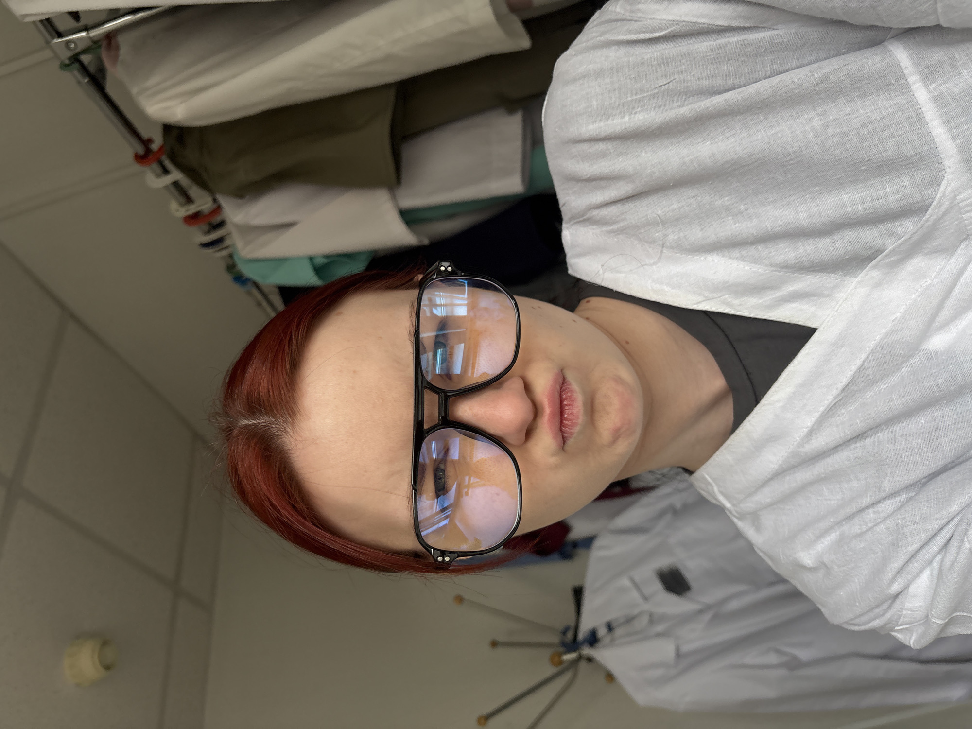 The image shows a close-up of a young woman with red hair wearing large, black-framed aviator glasses and a white lab coat. She is making a puckered-lip expression. In the background, there are several white lab coats hanging on a rack, and a ceiling light fixture is visible. The scene appears to be an indoor setting, possibly a hospital or laboratory. The specific location in Saint Petersburg, Russia is not discernible from the image.