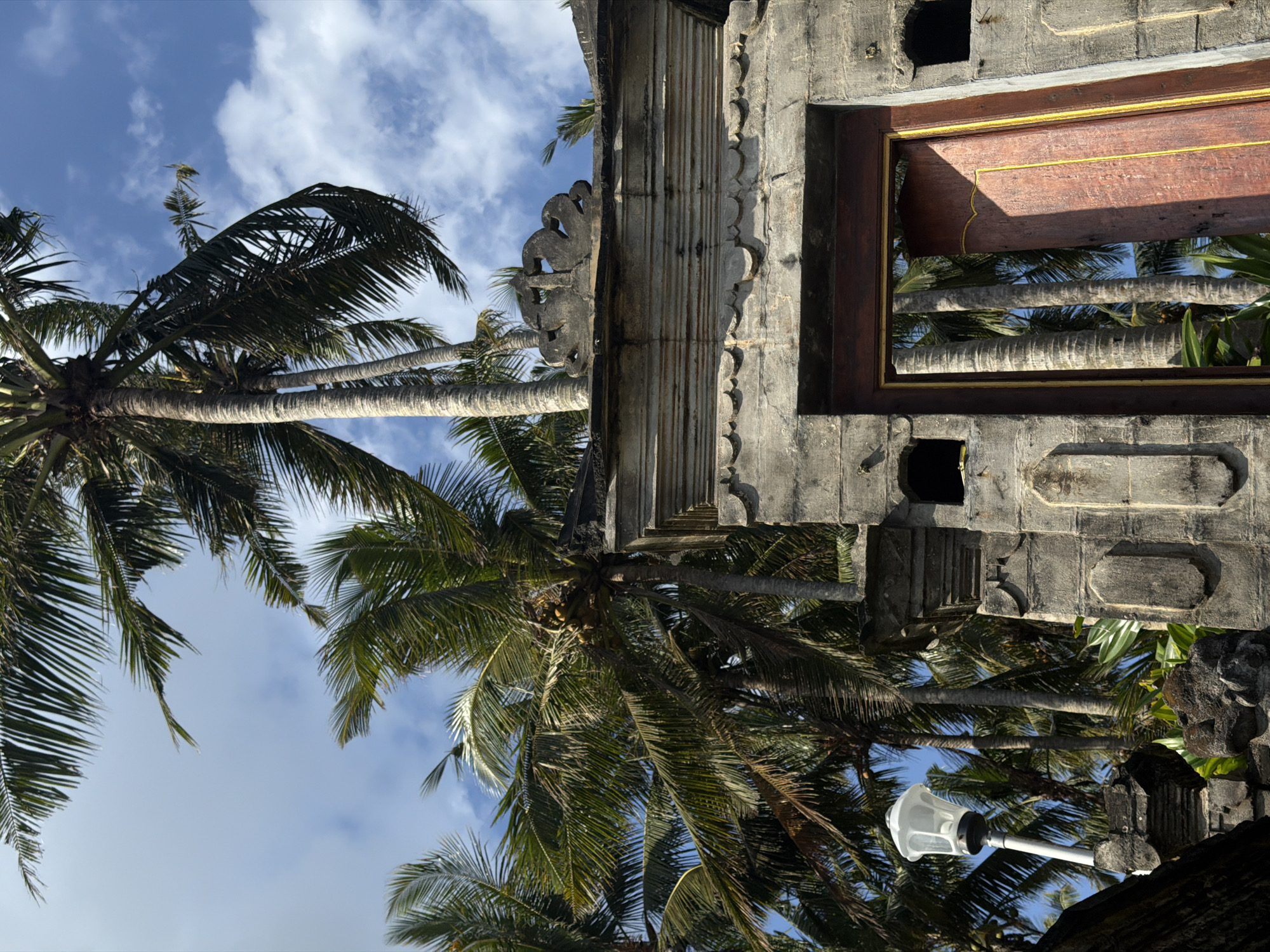 The image depicts a scene in Selemadeg Timur, Indonesia, featuring palm trees and a partially visible stone structure. The camera angle is low, looking upward toward the sky. The sky is blue with scattered clouds. Several mature palm trees with green fronds fill the foreground and left side of the frame. The stone structure, possibly a temple or gate, is on the right side and at the top of the image. It features carved details and a dark wooden element with a gold trim. A white street lamp is visible towards the bottom right. The image is rotated to the left 90 degrees.