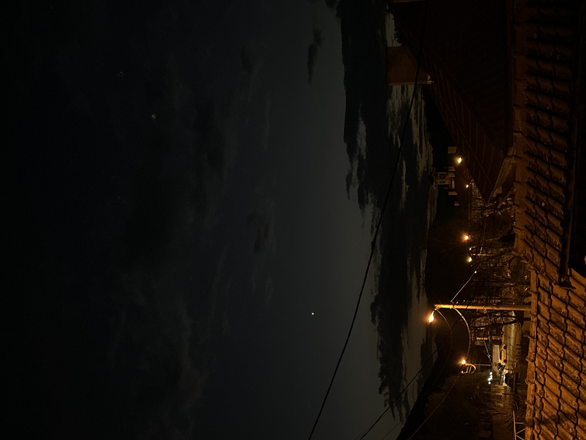 The image depicts a night scene in what appears to be a residential area. The sky is dark with clouds, and a single bright star or planet is visible. Streetlights illuminate the road, with a line of glowing bulbs extending into the distance. On the right side of the frame, the edge of a building with a tiled roof is visible. Power lines run across the scene. The location is unknown, with the city also being unknown.
