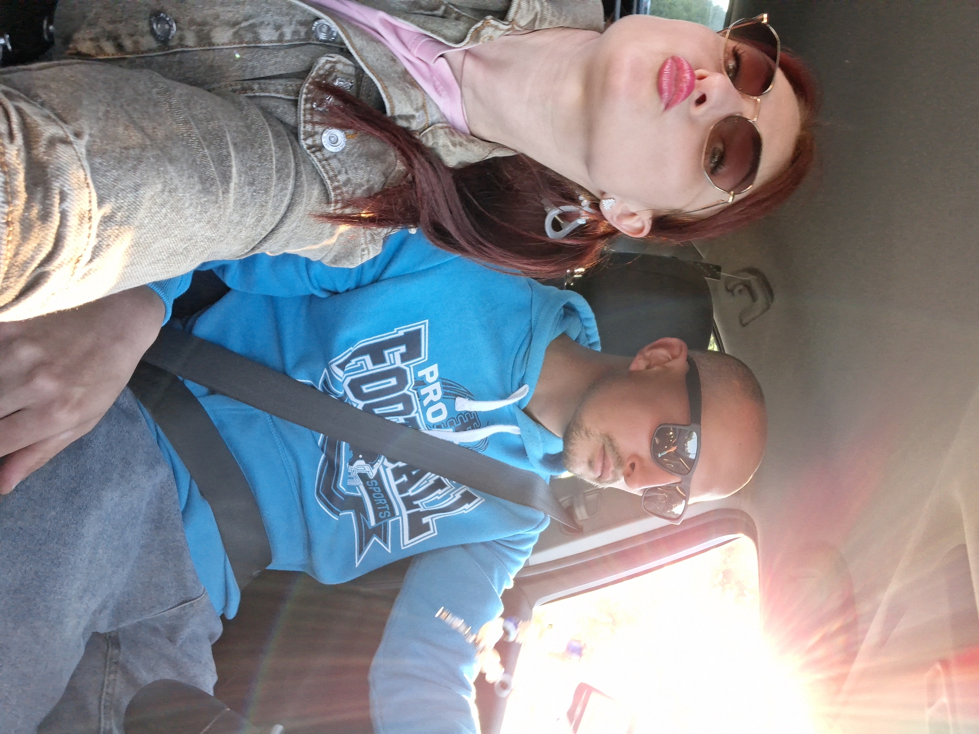 Two individuals, a woman and a man, are seated inside a car, likely in motion, as indicated by the angle of the sun and potential motion blur. The woman, on the left, has reddish-brown hair styled in a ponytail and is wearing large, hexagonal-shaped sunglasses and a denim jacket over a pink shirt. She is puckering her lips for a selfie. The man, on the right, is bald and wearing sunglasses. He is sporting a blue hoodie with "PRO FOOTBALL SPORTS" printed on it and is wearing a seatbelt. The interior of the car is visible, including the dashboard and headliner. The bright sunlight is streaming in through the windshield, creating lens flare. The location is estimated to be in or near Смалявічы, Belarus, given the provided context.