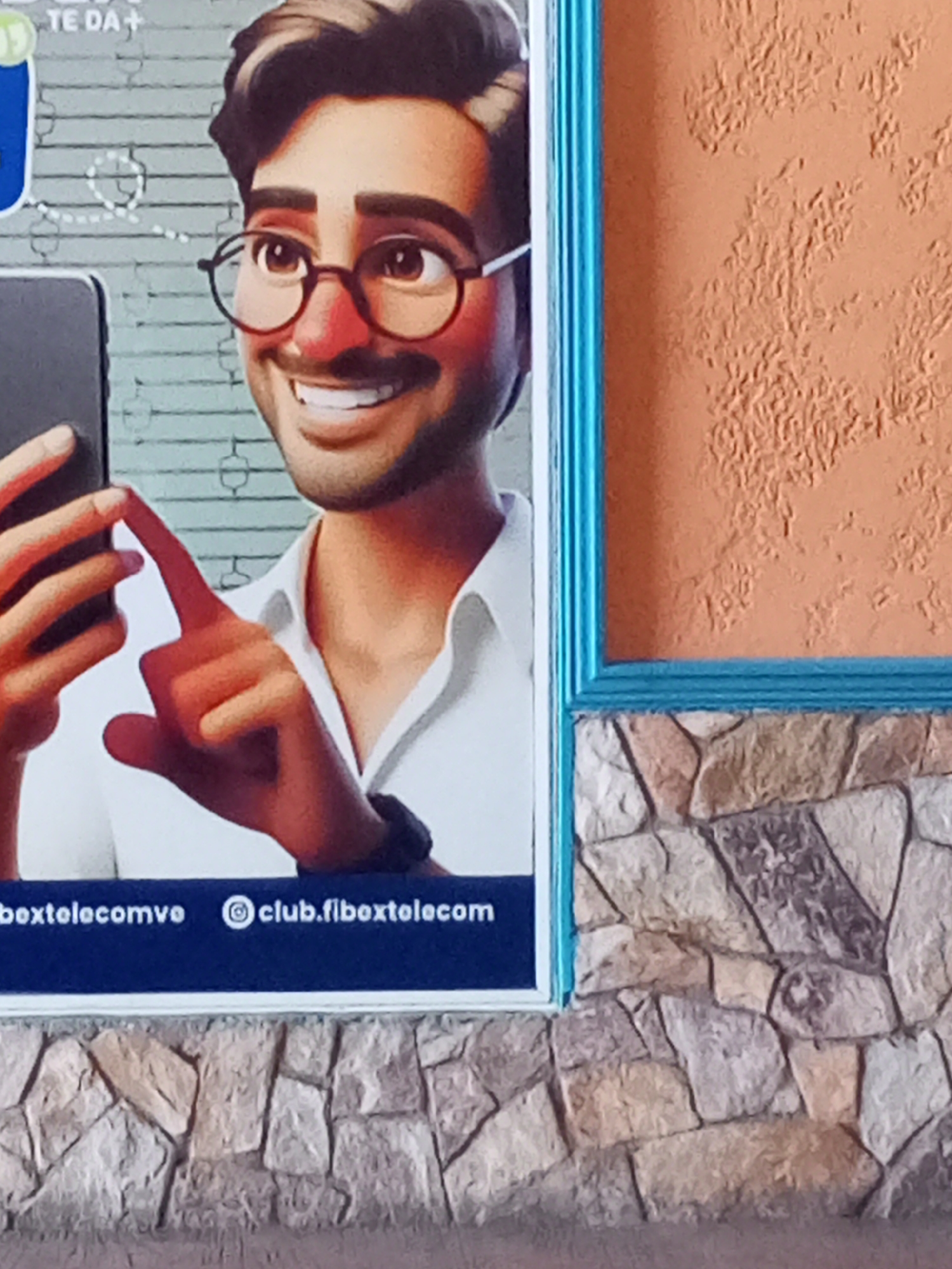 The image shows a close-up of a billboard featuring a cartoonish depiction of a man wearing glasses and a white shirt, holding a smartphone. The billboard has a blue border and is positioned against a textured orange wall and a stone wall at the bottom. The text "boxtelecomve" and "club.fibxtelecom" is visible at the bottom of the billboard. The man's skin tone is tan, and he has dark hair and a mustache. His eyes are brown, and he is smiling. His right hand is extended, and his index finger is pointing towards the smartphone. The billboard appears to be located in an outdoor urban setting, possibly on a building facade in Juan Griego, Venezuela, indicated by the presence of local text on the billboard. The overall scene is brightly lit, suggesting daytime.