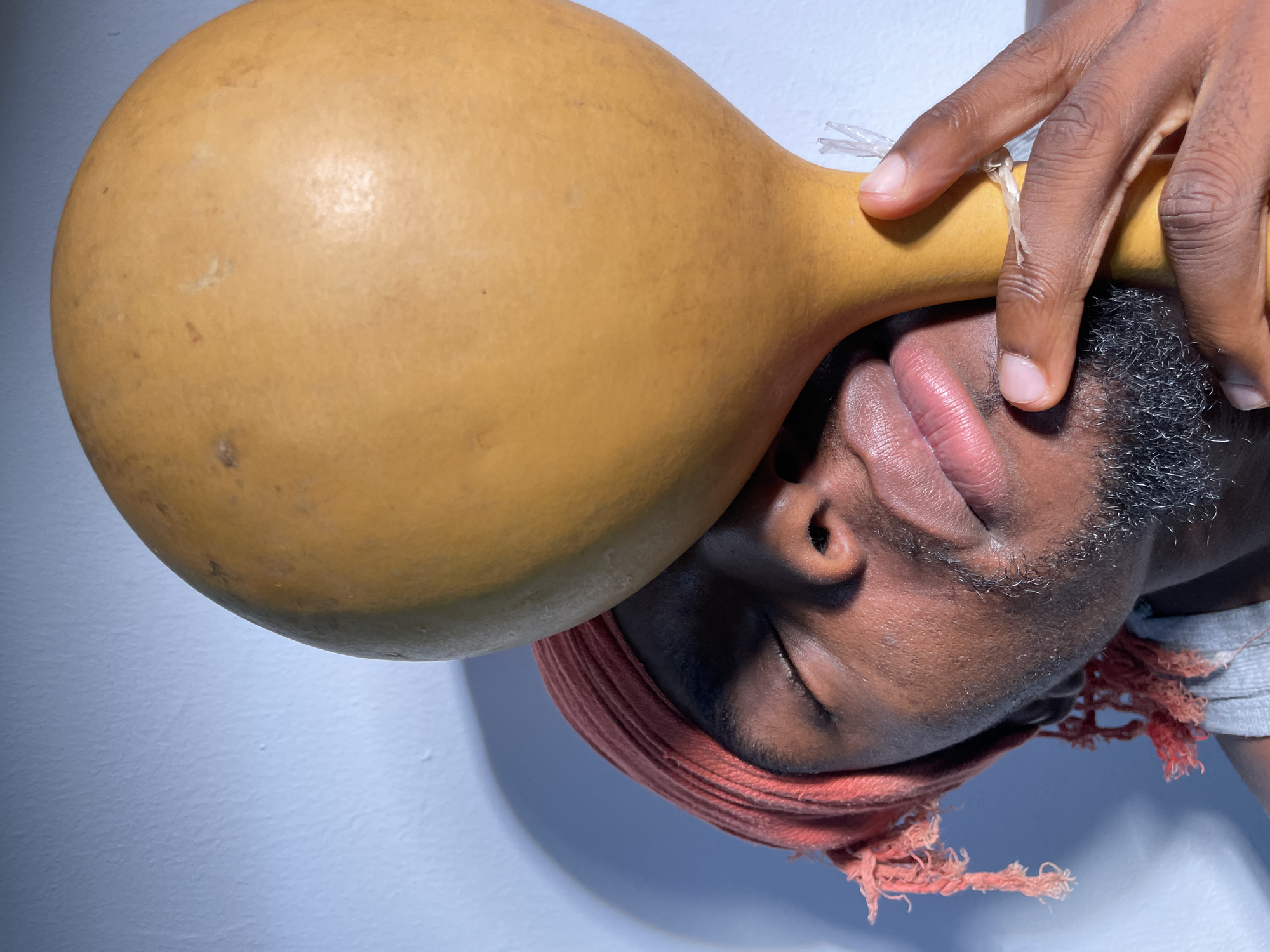 A person lies on their side, head wrapped in an orange cloth, holding a large, gourd-like object against their forehead. The gourd is yellow-tan with slight imperfections. The person's skin is dark, and their fingers are touching the gourd near a string-like attachment. The background is a light blue-grey. There is no mention of Barakin Akawo, Nigeria, in the prompt or available evidence.