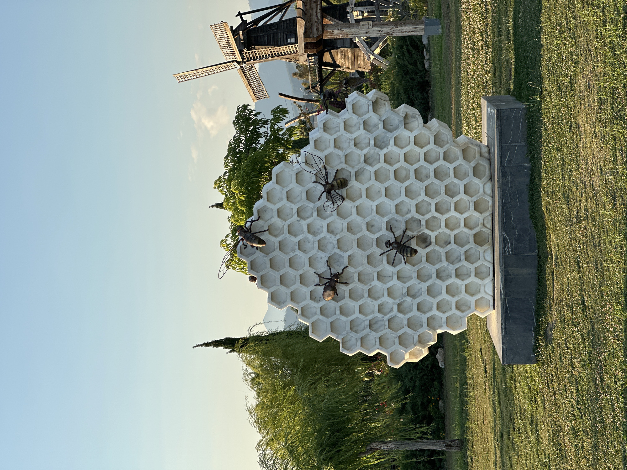 This image, set in Fethiye, Turkey, captures a serene outdoor scene centered around an artistic sculpture. A prominent, large hexagonal honeycomb, crafted from a light-colored, possibly marble, material, is positioned in the foreground. Several metallic bee sculptures are meticulously detailed and affixed to the honeycomb's surface, enhancing its realistic appearance. The entire sculpture is mounted on a dark, rectangular pedestal with subtle marble veining.
In the middle ground, an old wooden windmill with large sails stands against a clear blue sky, hinting at distant mountains. The area is rich with vibrant greenery, including leafy trees, shrubs, and a tall, slender conifer on the left. The ground is covered in well-maintained green grass, displaying long shadows that indicate either early morning or late afternoon sunlight, casting a warm glow across the landscape. The sky is largely clear, with minimal cloud cover. No specific activity is taking place, and no people or visible text are present, contributing to a tranquil and picturesque ambiance.