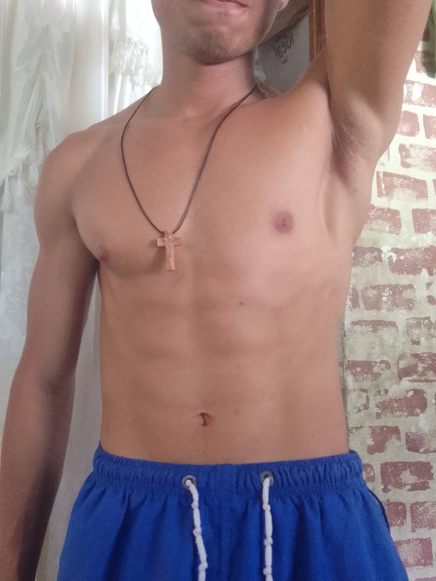 A bare-chested man is seen from the upper thighs up. He is wearing blue shorts with a white drawstring. He has a wooden cross necklace. He is indoors; a brick wall is visible behind him. A white curtain is also visible to the left. The location is Juan Griego, Venezuela.
