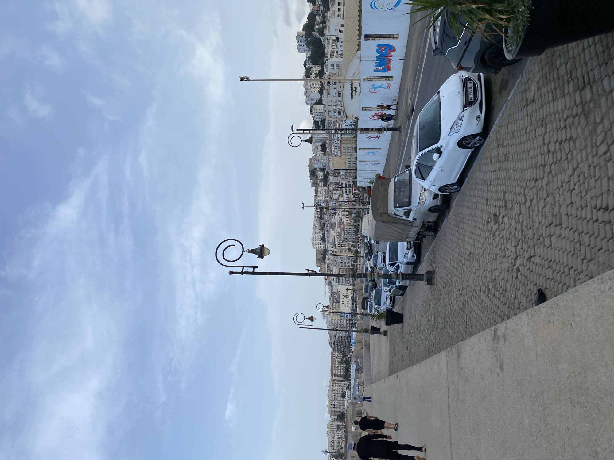 The image captures a street scene in Beau Fraisier, Algeria, with a view of a coastal city. In the foreground, a concrete walkway runs along the left side of the image, with two figures walking away from the camera. The walkway is bordered by a cobblestone street where several cars are parked, including a white Peugeot and a dark SUV. A light-colored truck is also visible.
The street is lined with decorative lampposts featuring a spiral design. In the background, a dense cityscape of buildings with white facades stretches up a hillside. A white wall adorned with colorful graffiti, featuring abstract shapes and text, runs along the side of the street. The sky is overcast with a pale blue hue and scattered clouds. The overall atmosphere suggests an urban coastal environment.
