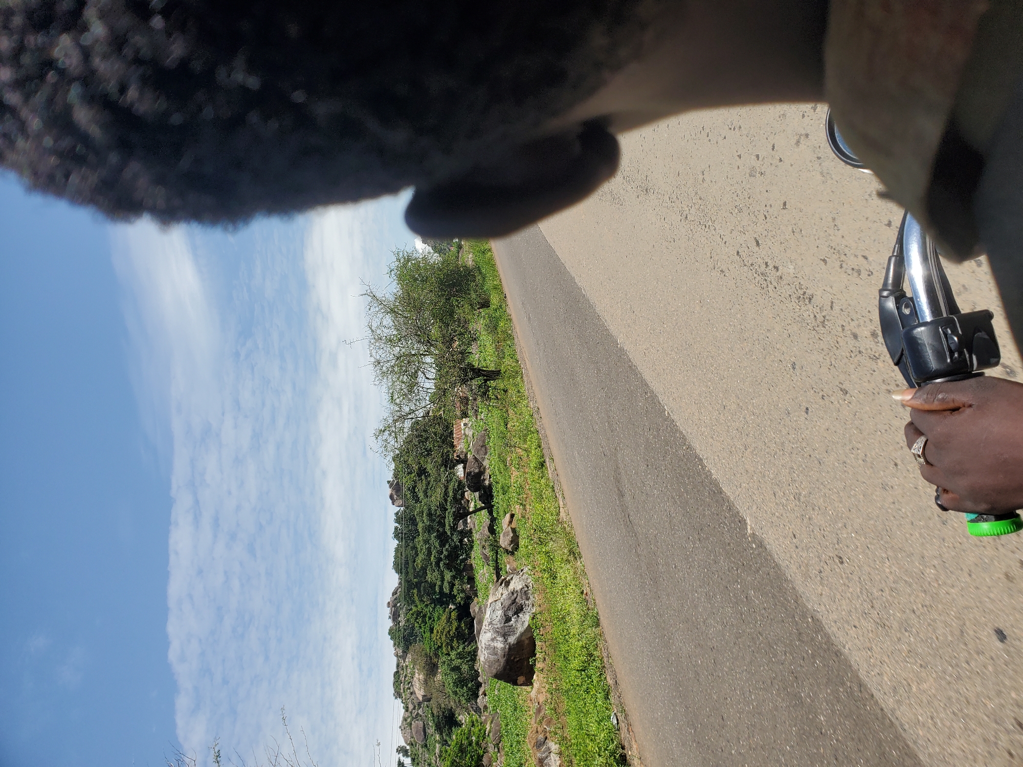 The image shows a person's head and shoulder in the foreground, looking forward. The person is riding a motorcycle on a paved road. The road curves to the left and is bordered by a grassy area with rocks and trees on the right. In the background, there are more trees and rocky hills. The sky is blue with some white clouds. The scene appears to be in a rural or semi-rural area. The overall impression is one of travel and observation of the surrounding landscape.