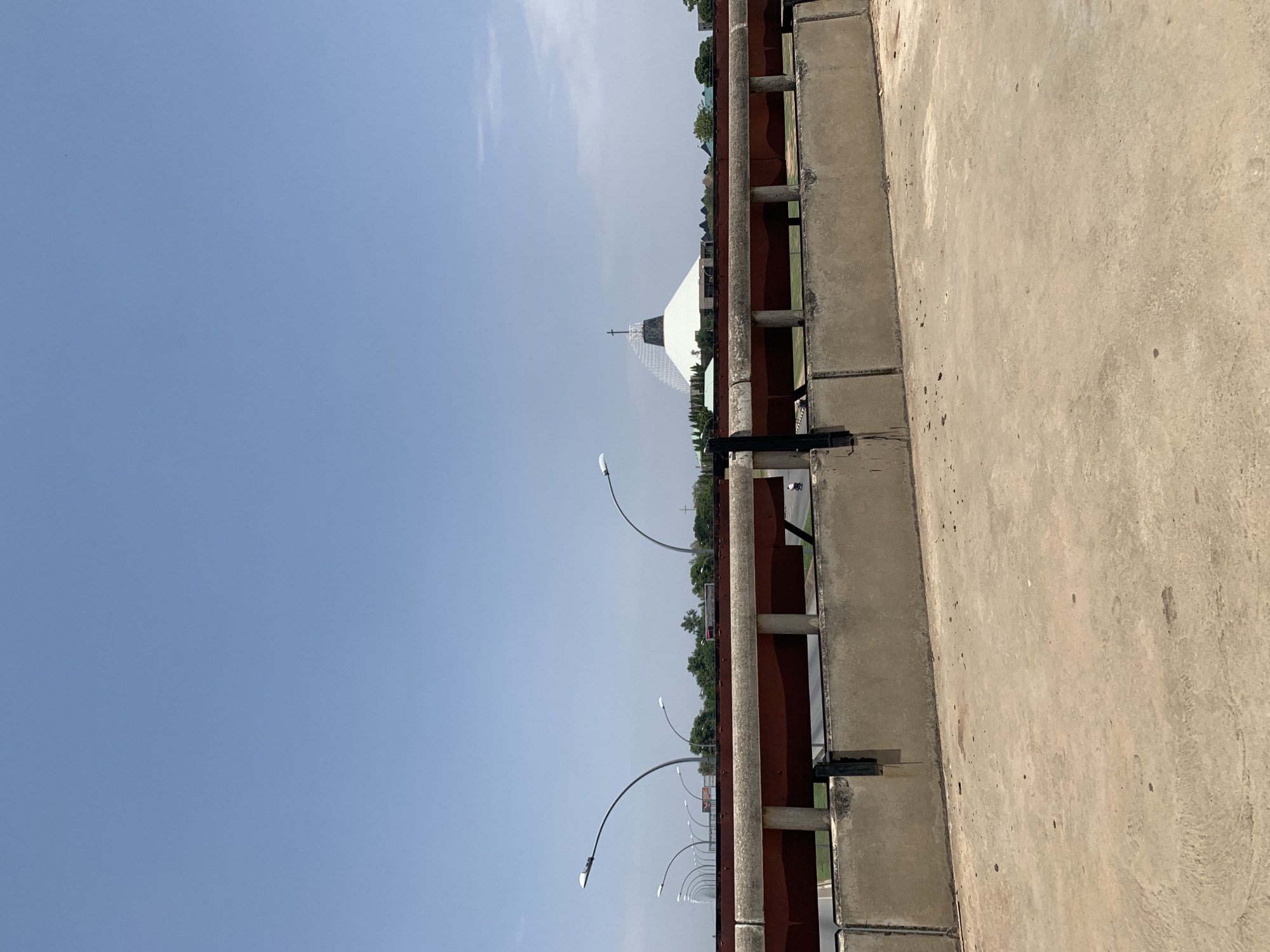 The image shows a view from a bridge or overpass in Kubwa, Nigeria. The scene features a weathered concrete barrier with a rusted metal railing. Beyond the railing, there is a view of a multi-lane road with streetlights and sparse traffic. In the distance, there's a large, white, pyramidal structure with a cross on top, likely a church or religious building. The sky is clear and pale blue. The environment appears urban, and the perspective is from a stationary, elevated position.