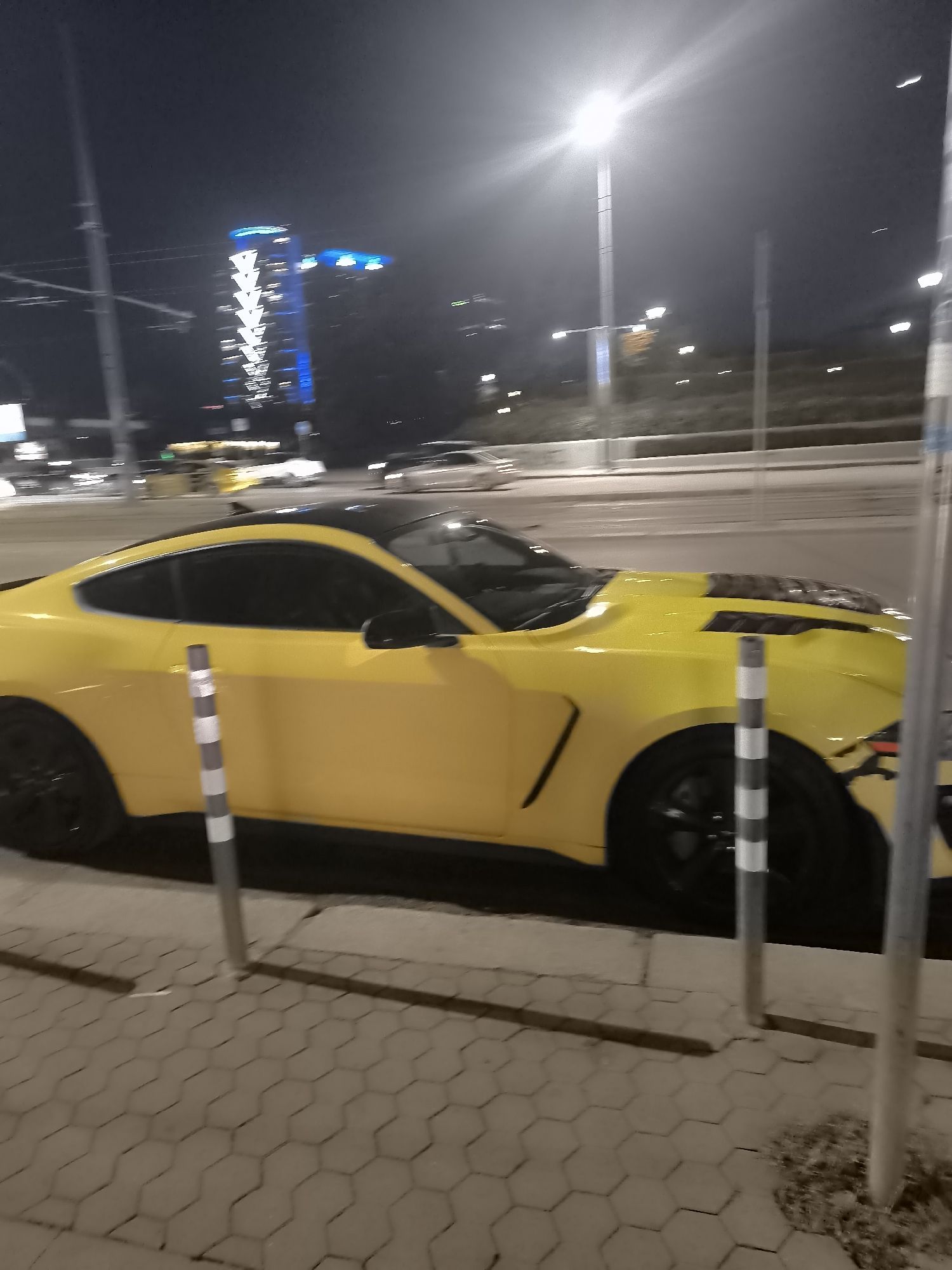 A nighttime scene shows a yellow Ford Mustang parked on a cobblestone sidewalk in Sofia, Bulgaria. The car has a black roof and hood, and black wheels. In the background, there is a tall building with blue lights. Street lights are visible, and other cars are on the road. Three black and white striped bollards are located close to the curb.