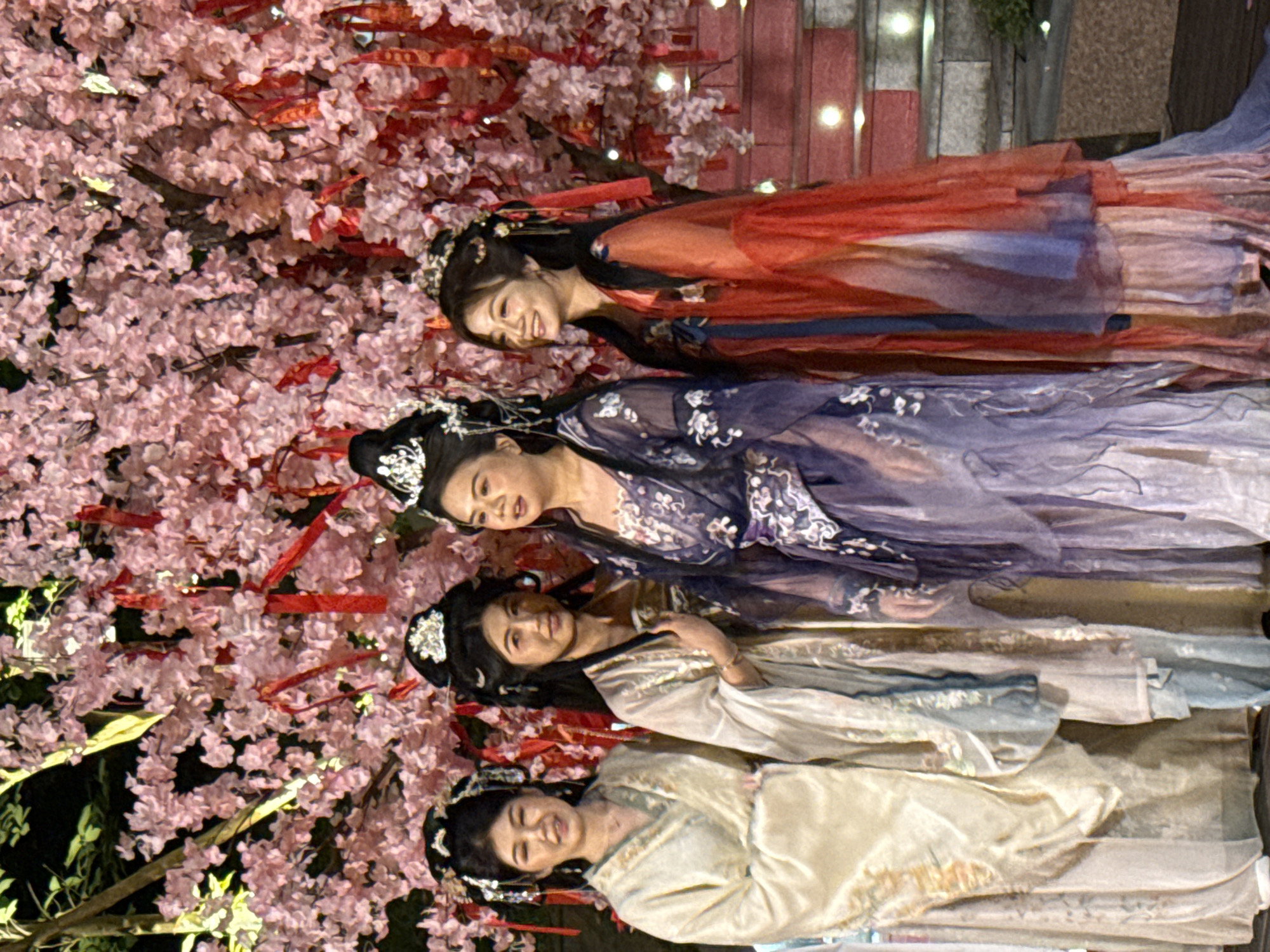 Four women dressed in traditional Chinese clothing stand next to a flowering tree with red ribbons in Jiulongpo District, China. The women are arranged in a vertical line, each wearing a different color of clothing: red, purple, light blue, and tan. All have traditional Chinese hairstyles with jeweled accessories. The flowering tree is on the left side of the image and has pink blossoms and several red ribbons tied to its branches. A set of stairs is visible in the background. The scene is lit with artificial lighting, creating a bright and somewhat surreal atmosphere.