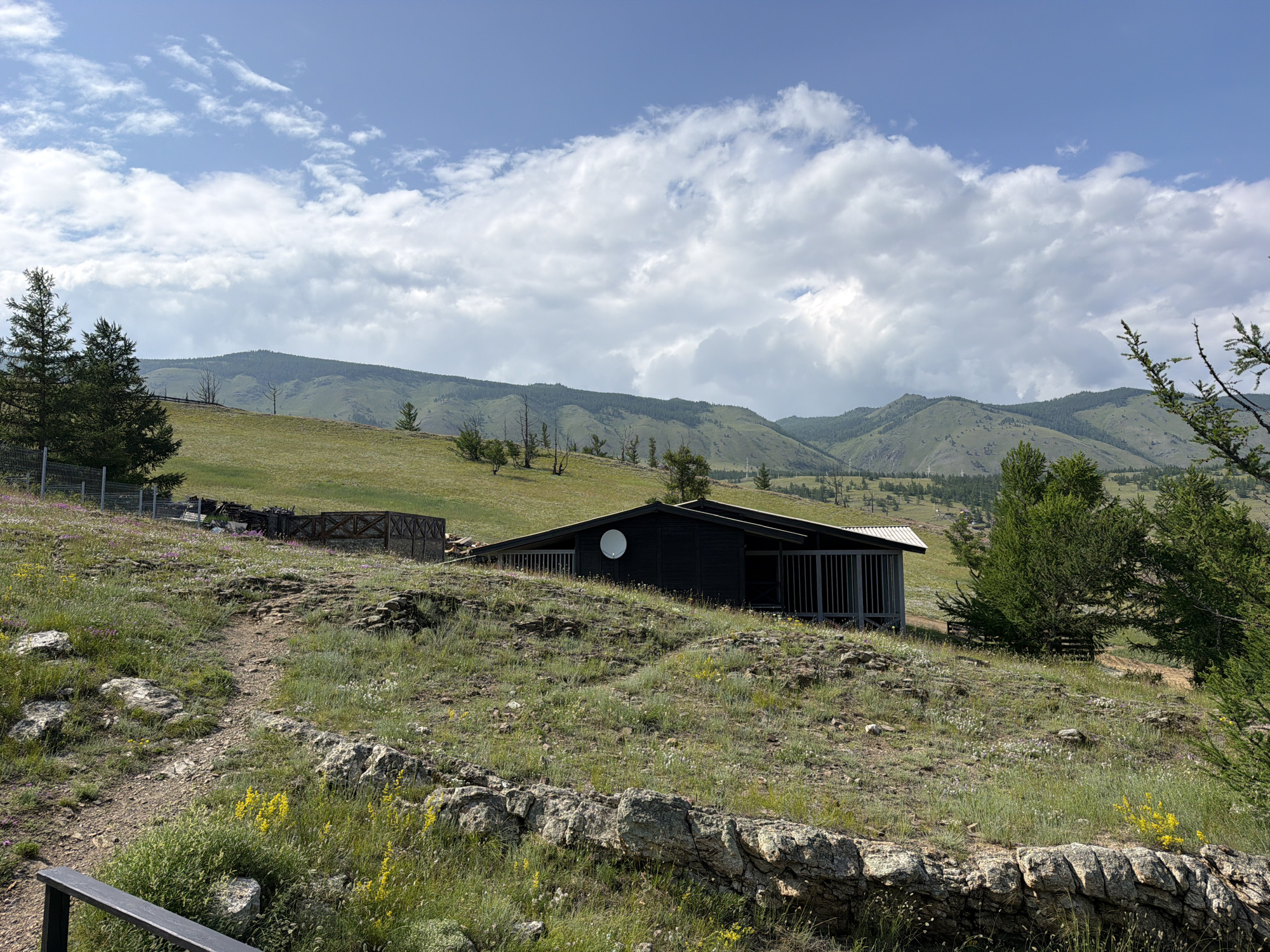 The image depicts a rustic scene in the village of Sarma, Russia. In the foreground, a rocky, grassy hillside is dotted with small yellow wildflowers. A dirt path winds its way up the hill. In the midground, a dark, modern-looking house with a white satellite dish is situated on a grassy slope. The house has a covered porch with vertical gray slats. Beyond the house, a wooden fence encloses a fenced area with some stacked wooden materials. Rolling green hills and mountains, some covered in dark forests, form the backdrop under a partly cloudy sky. Several evergreen trees are scattered across the landscape.