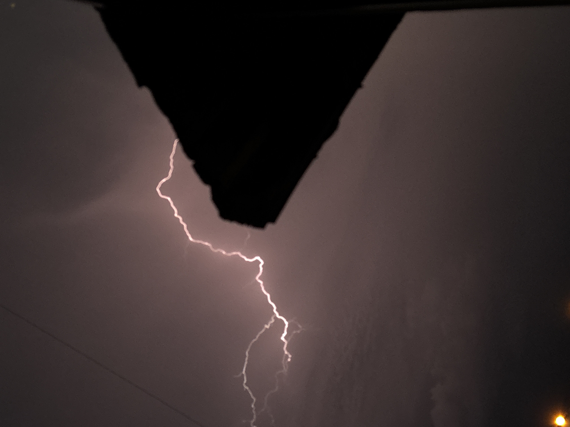 The image captures a lightning strike during a storm. The scene is dark, likely at night. A dark, triangular object, possibly part of a building, is in the upper portion of the frame. A jagged lightning bolt is visible against the cloudy sky. There are no people visible. The bottom right corner has a faint blurred light source, potentially a streetlamp. The presence of powerlines on the left side indicates an urban environment, such as Varna, Bulgaria, where power lines are part of the city infrastructure.