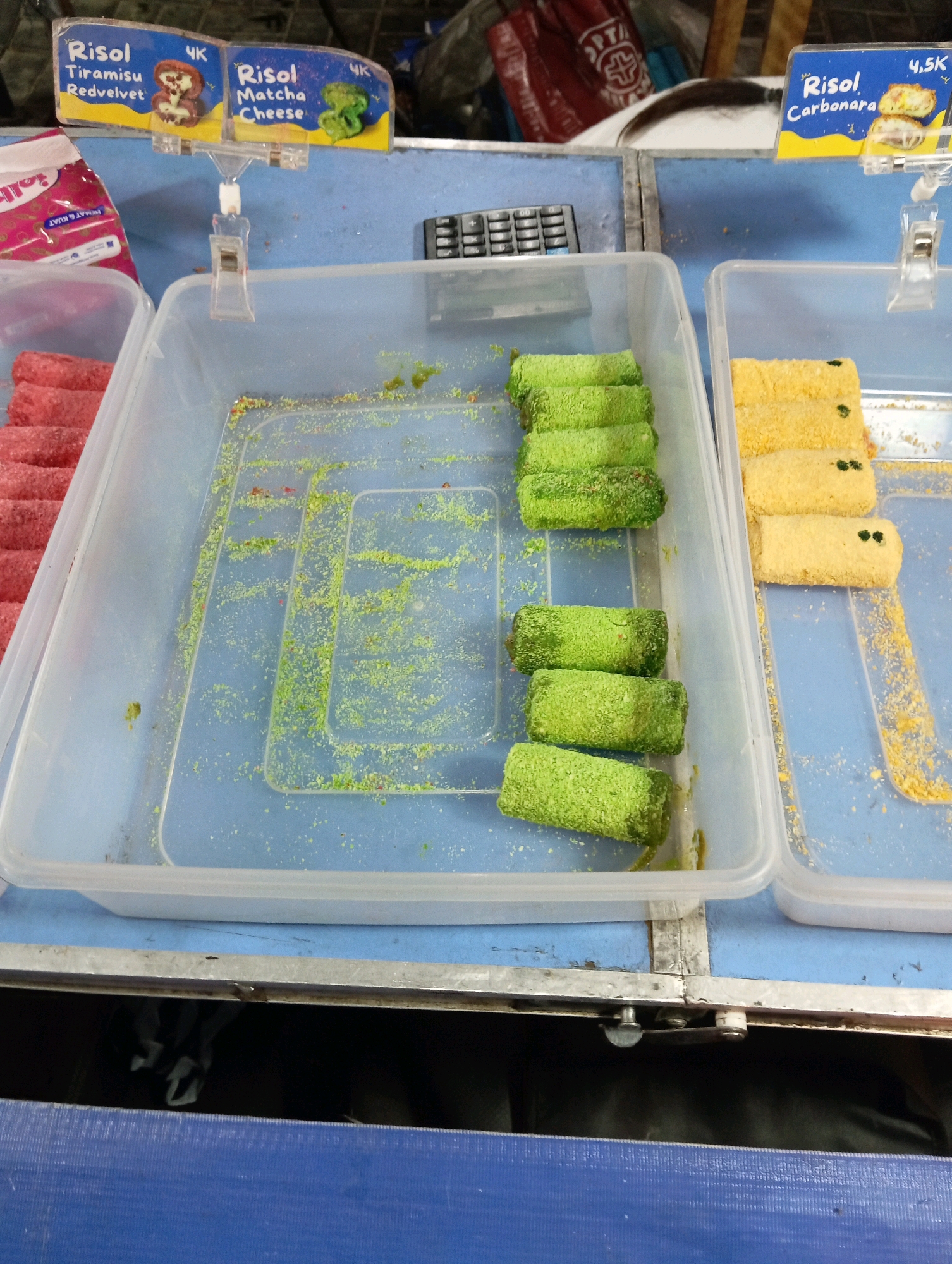 This image depicts a food stall in Magelang, Indonesia, likely at a local market or street food vendor. In the foreground, several plastic containers are filled with food items. One container holds several bright green, rolled pastries, possibly filled with matcha or a similar green filling, covered in a fine green crumb coating. Scattered around this container and on the blue surface are small green and red sprinkles. To the right, another container holds yellow rolled pastries, also coated in crumbs, with some small dark green pieces visible on top. In the background, a red bag with white text is partially visible. Above the containers, two signs are attached to a metal rack. The left sign, with a blue background, advertises "Risol Tiramisu Redvelvet" and "Risol Matcha Cheese," each with a small illustrative image. The right sign, also blue, advertises "Risol Carbonara" and shows a picture of the pastry cut open. The overall environment appears to be an outdoor or semi-outdoor stall, with a blue surface dominating the scene and hints of other market goods in the background.