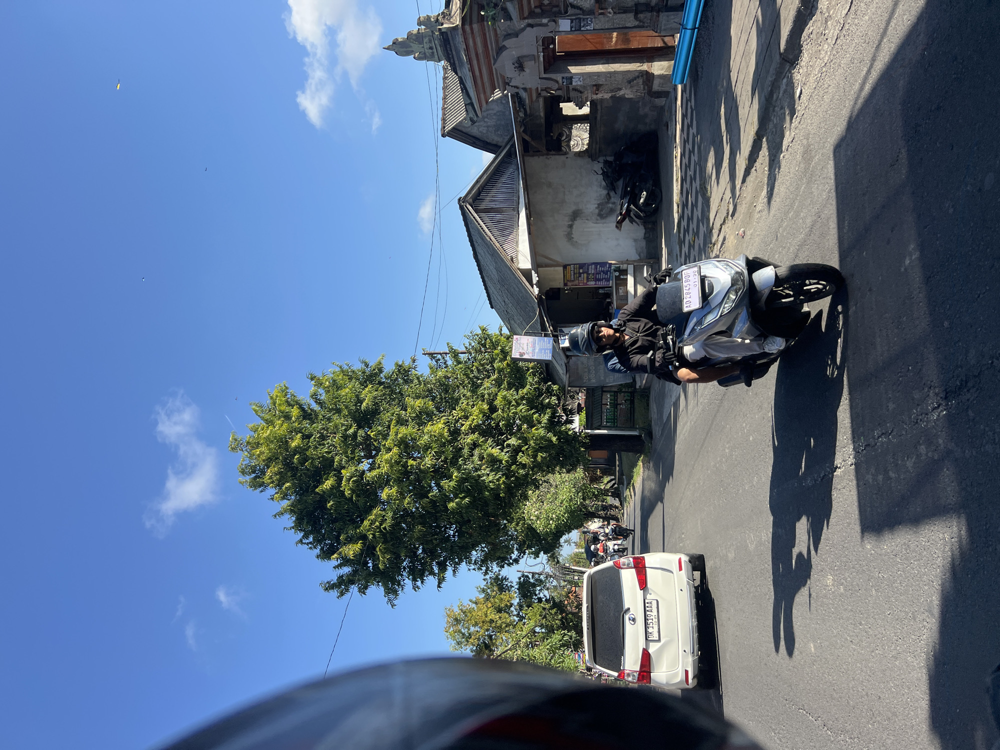 A perspective shot from a moving vehicle in Sukawati, Indonesia, shows a street scene under a clear blue sky with scattered clouds. In the foreground, the dark helmet of the photographer's vehicle is partially visible on the left. The road ahead is asphalt, with a white SUV in the middle lane, its license plate reading "DK 3519 AAA." To the right, a person wearing a black jacket and helmet rides a silver motorcycle, their license plate displaying "AD 2845 BQF." The rider is looking forward, and the motorcycle is in motion.
The right side of the street features a building with a gray, textured facade and a grey tiled roof. A wooden door and intricately carved stone archway are visible. Further down the street, more buildings with tiled roofs line the road. A tall, leafy green tree stands prominently on the left side of the street, casting a shadow on the road. Other trees and vegetation are present along the sides of the street, with power lines overhead. In the distance, other motorcycles and a vehicle can be seen on the road. The overall atmosphere is that of a bright, sunny day in a bustling Indonesian town.