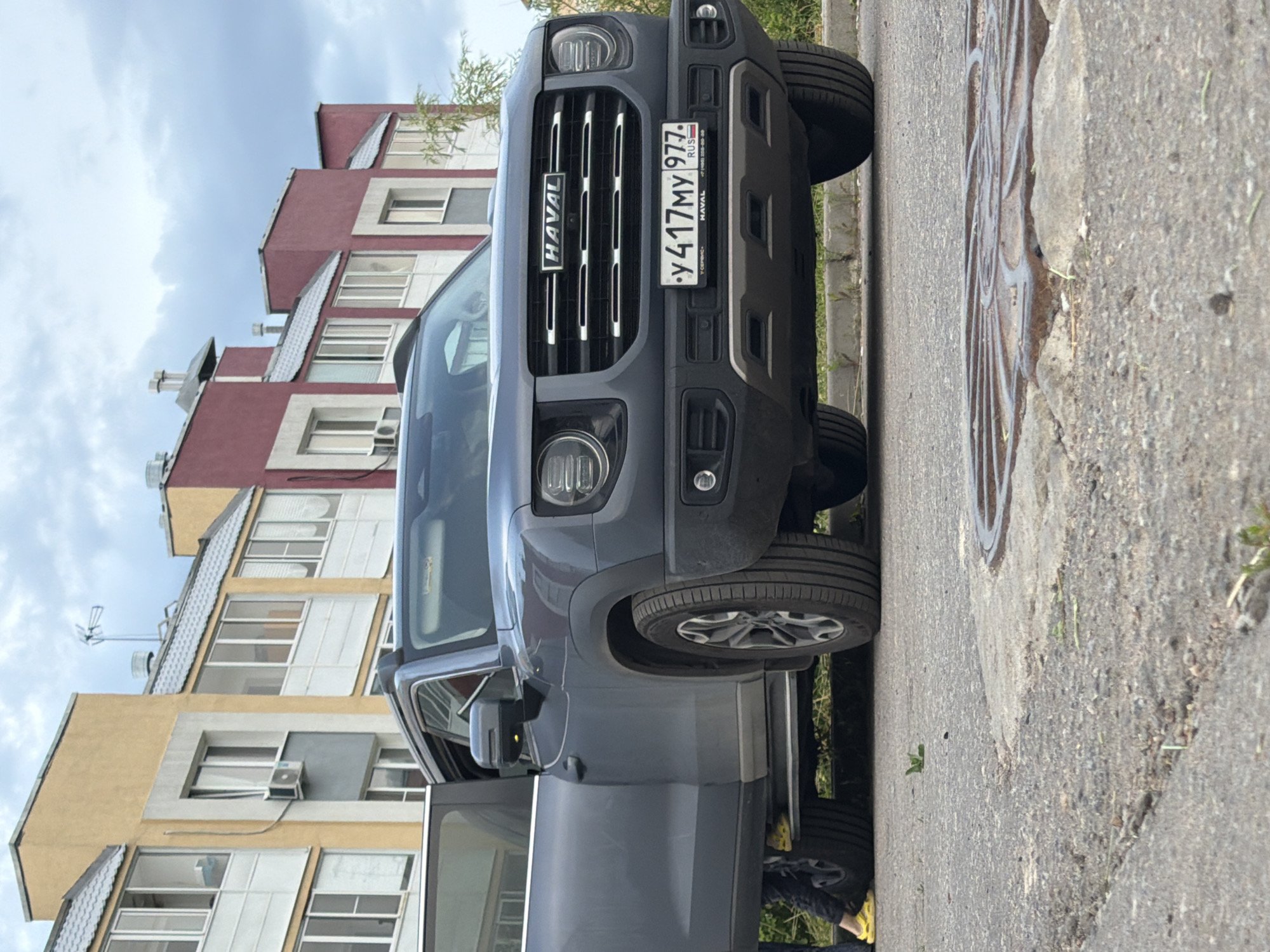 A dark gray Haval SUV is parked on a paved road in front of a row of colorful buildings. The driver's side door of the SUV is open. A person wearing yellow shoes and dark pants is partially visible standing next to the open door. The SUV has a license plate that reads "y 417MY 9977 RUS". The buildings in the background are multi-story with yellow, red, and beige facades, featuring numerous windows and visible air conditioning units. The sky is overcast with clouds. A manhole cover is visible on the road in the foreground. The overall scene appears to be in a residential urban environment.