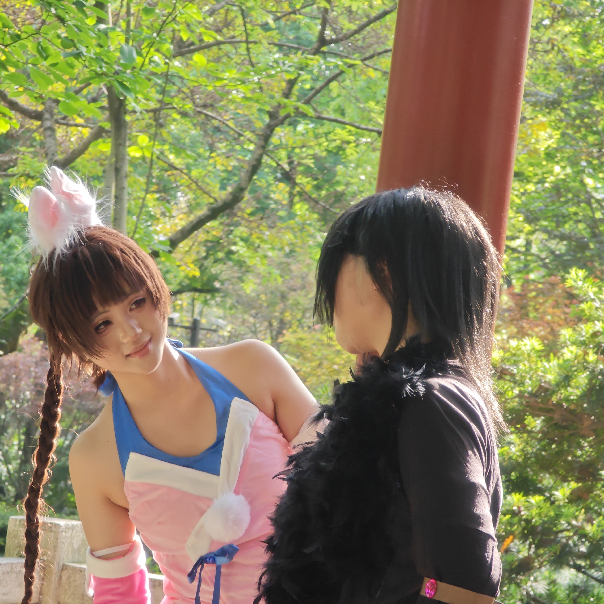 The image depicts two individuals in cosplay attire in an outdoor setting, possibly a park or garden. The location is the city of Yuzhong District, China.
The individual on the left is dressed in a pink and blue outfit resembling a bunny costume, complete with bunny ears. They have brown hair styled with a braid and are looking towards the other individual.
The individual on the right is wearing a black outfit with a black feather boa. Their hair is black and obscures part of their face.
The background includes trees and foliage, suggesting a natural environment. A red pillar can also be seen, indicating a structure like a pavilion or temple.