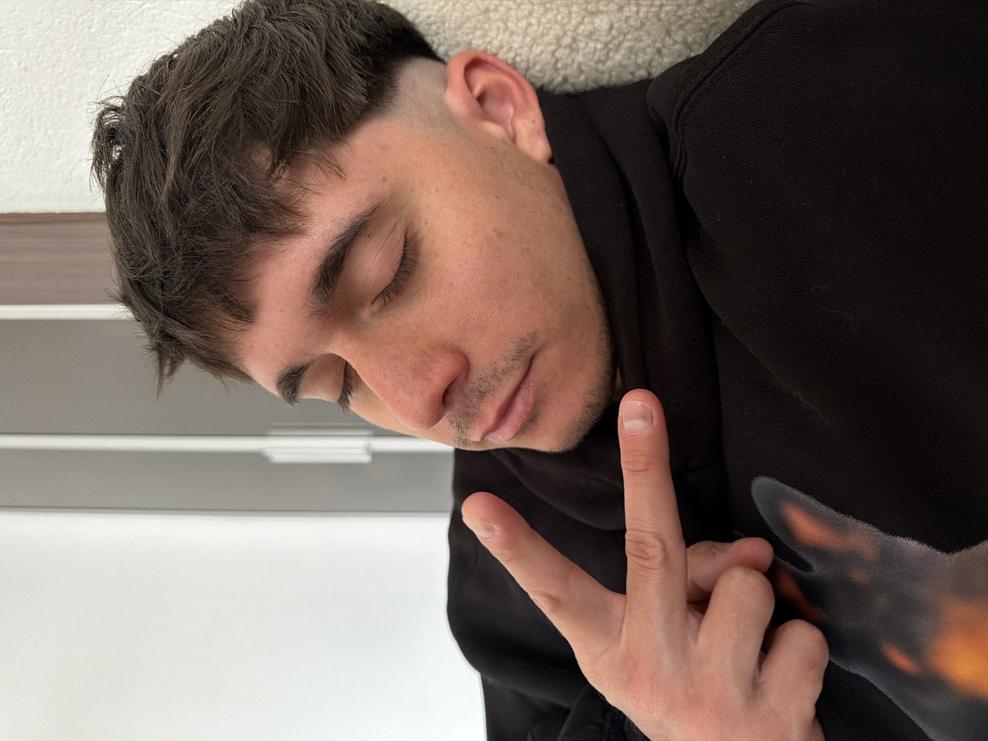 An eye-level shot captures a young man in a relaxed, indoor setting, possibly in Blagoevgrad, Bulgaria. He is positioned diagonally across the frame, with his head tilted downwards and eyes closed, suggesting he is resting or asleep. He has dark, wavy hair with a closely shaved fade on the sides, and light stubble on his chin and upper lip. He wears a black long-sleeved garment, possibly a sweatshirt or hoodie, which features a subtle, glowing graphic of what appears to be a dog's head, resembling a Doberman, on his right side. His right hand is raised in front of his chest, forming a peace sign with his index and middle fingers. The background is simple, consisting of a textured light-colored surface behind his head and a plain white or light grey wall in the lower left portion of the image. The overall impression is one of peaceful repose, with a casual gesture.
