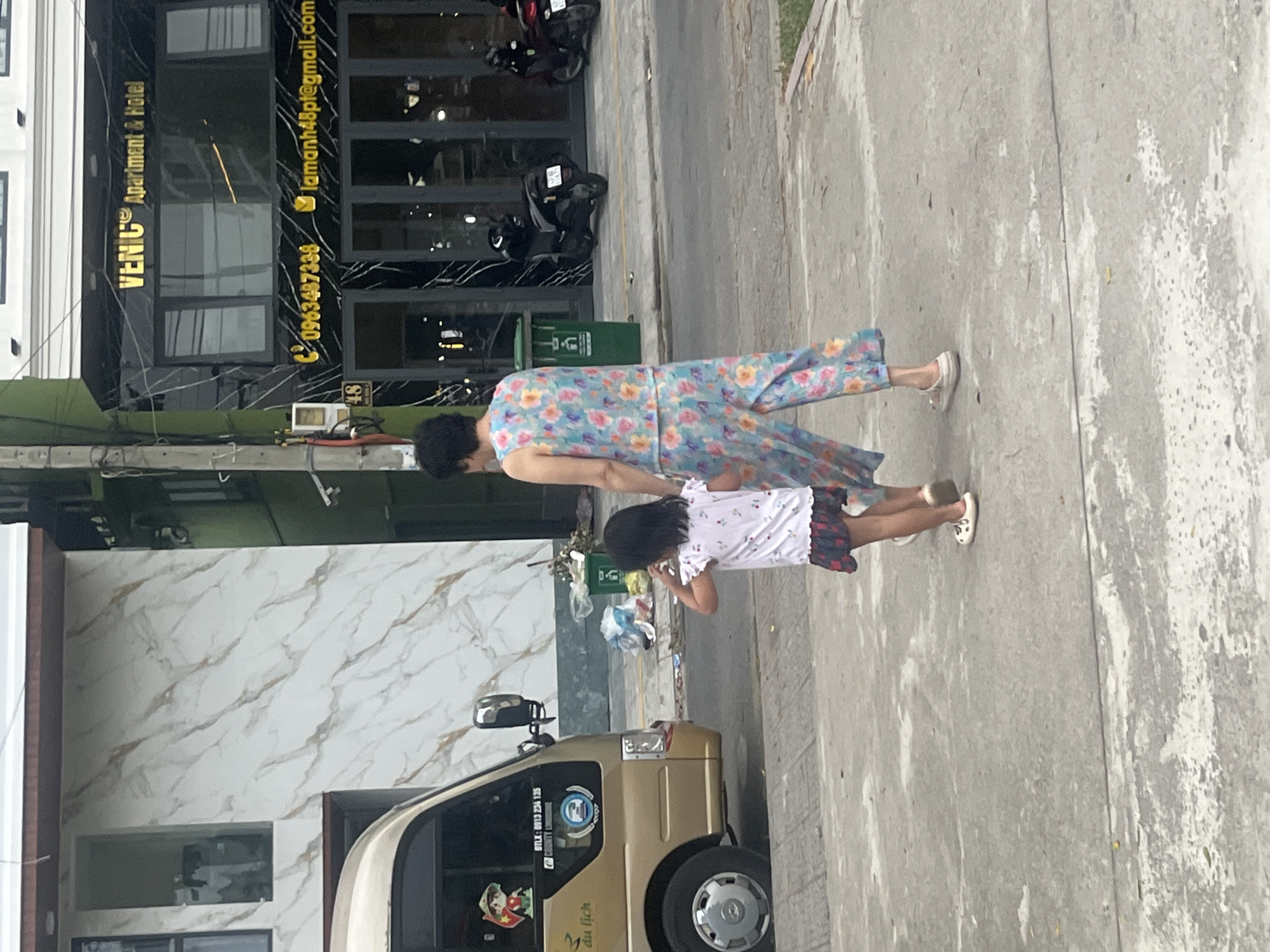 A woman and a young girl are walking on a sidewalk in front of a building. The woman is wearing a floral print jumpsuit, and the girl is wearing a white t-shirt and a skirt. A gold van is parked on the street to the left of the sidewalk. The building behind them has a black facade with gold lettering that reads "VENIC Apartment & Hotel." The street appears to be in Đà Nẵng, Vietnam, indicated by the text on the van.