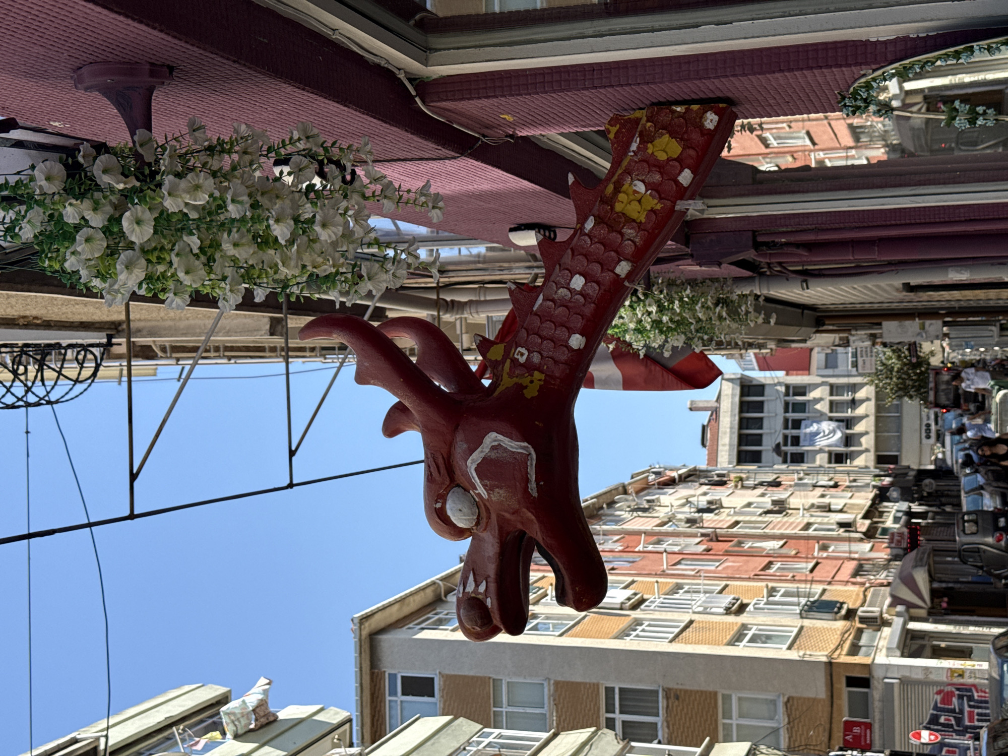 The image shows a close-up of a red, stylized dragon head ornament mounted on a building facade in Kadıköy, Turkey. The dragon's head is detailed with white eyes and a slightly open mouth. Its body, adorned with red scales and yellow and white accents, extends to the right, partially obscuring a Danish flag. Above the dragon, a cluster of white artificial flowers hangs from a planter. The building itself has a textured, dark pink facade. In the background, a narrow street is visible, lined with multi-story buildings in various colors, including orange, red, and beige. Several windows, air conditioning units, and laundry hanging from balconies are apparent. The sky is a clear blue. The scene captures a narrow street in an urban environment with a distinct decorative element.