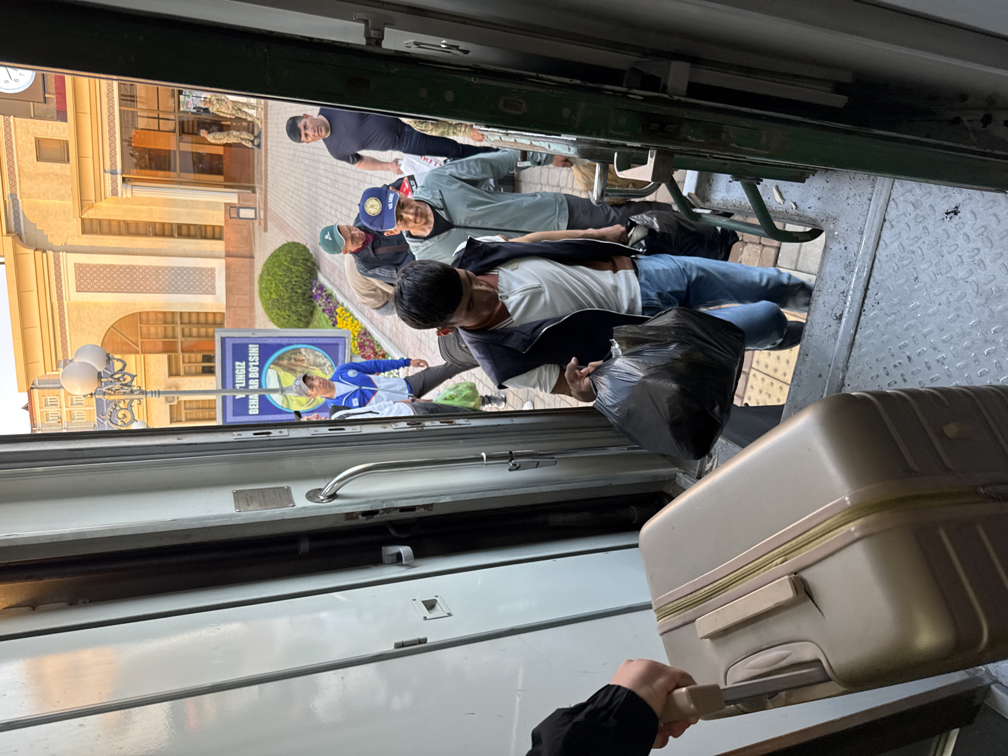 The image, taken from inside a train, shows a group of people on the train platform in Jizzakh, Uzbekistan. A person, presumably a passenger, is pulling a beige suitcase with wheels and an extendable handle into the train. The person is also holding a black plastic bag. Outside the train, several people are standing, some appearing to be waiting to board or disembark. One man is wearing a black t-shirt. Another man is wearing a blue baseball cap. A third man wears a blue baseball cap. A fourth man is wearing a blue athletic jacket. A nearby building has intricate architectural details, including arches and latticework. A poster is visible. Streetlights with ornate bases are positioned along the sidewalk.