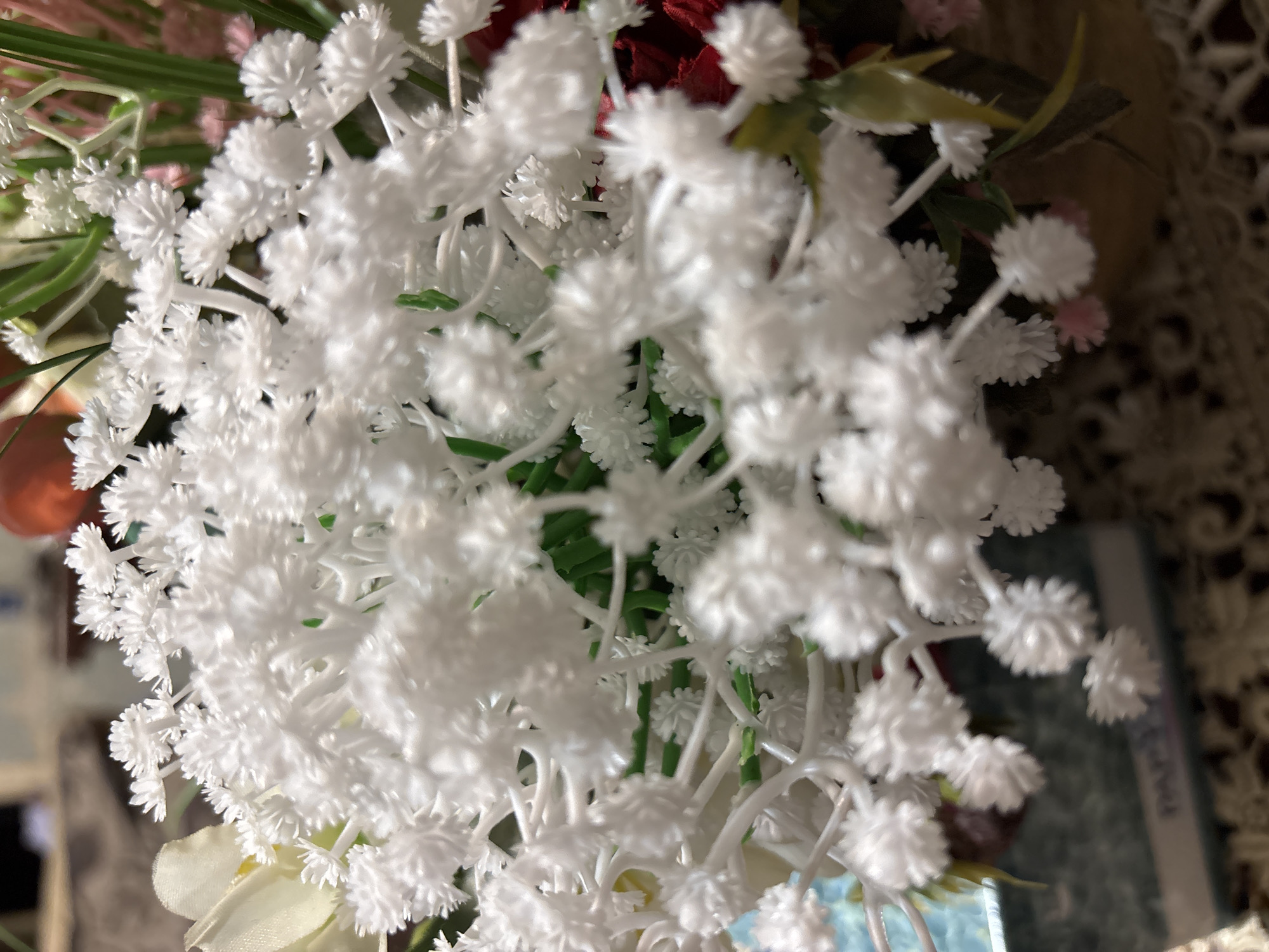 A close-up shot captures a vibrant arrangement of artificial flowers, dominated by a large cluster of small, white, spiky blossoms resembling baby's breath, positioned centrally and in the foreground. These white flowers appear to be made of plastic or a similar synthetic material, exhibiting a uniform texture.
The arrangement is densely packed, with several stems and individual white florets visible. Interspersed among the white flowers, green artificial foliage can be seen, providing a contrast in color. In the softly blurred background, hints of other artificial flowers are discernible, including dark red and light pink elements.
To the right, a cream-colored, intricate lace or crocheted pattern is visible, suggesting a doily or tablecloth, placing the setting likely indoors. Another object, possibly a light blue book or box with a white spine and faint, indecipherable text, is partially visible in the lower right background. The lighting is soft, creating a gentle illumination on the flowers, and a shallow depth of field keeps the foreground flowers in sharp focus while blurring the background. The overall impression is a decorative floral display in a home or similar indoor environment in Vagharshapat, Armenia.
